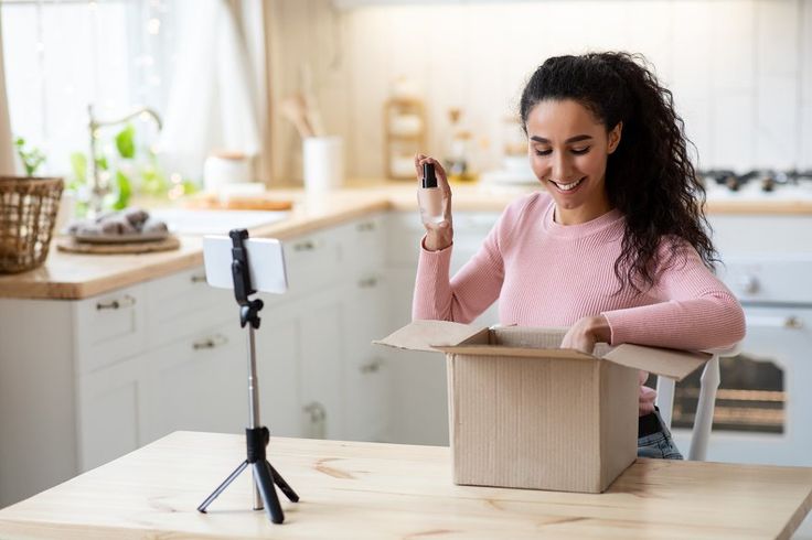 A smiling woman with dark curly hair records an unboxing video in a bright kitchen while holding a cosmetic product. She is seated at a wooden table with a smartphone mounted on a tripod, capturing her reaction as she reaches into a cardboard shipping box.