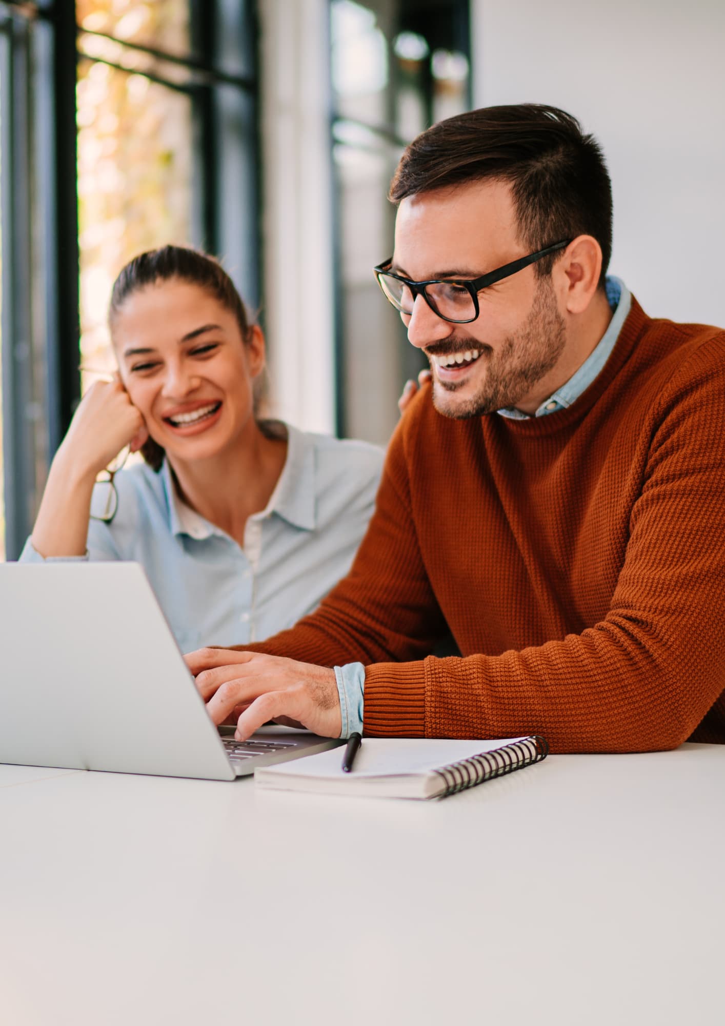 A smiling man with glasses wearing an orange sweater types on a laptop while a woman sitting next to him laughs and looks at the screen in a bright office or home workspace.