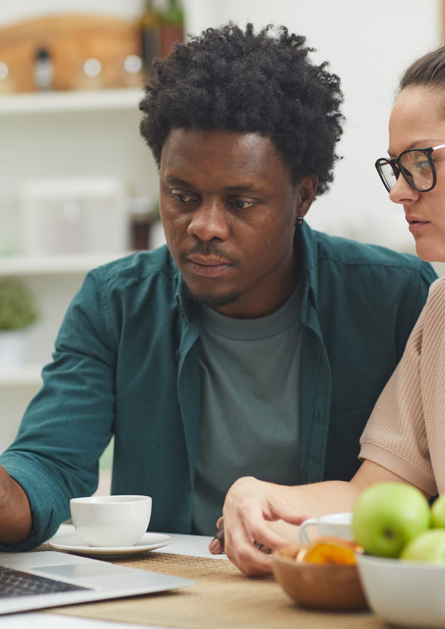 A focused Black man with an afro hairstyle and a green button-down shirt looks intently at a laptop screen alongside a woman wearing glasses, sitting at a kitchen table with coffee cups and a bowl of green apples.