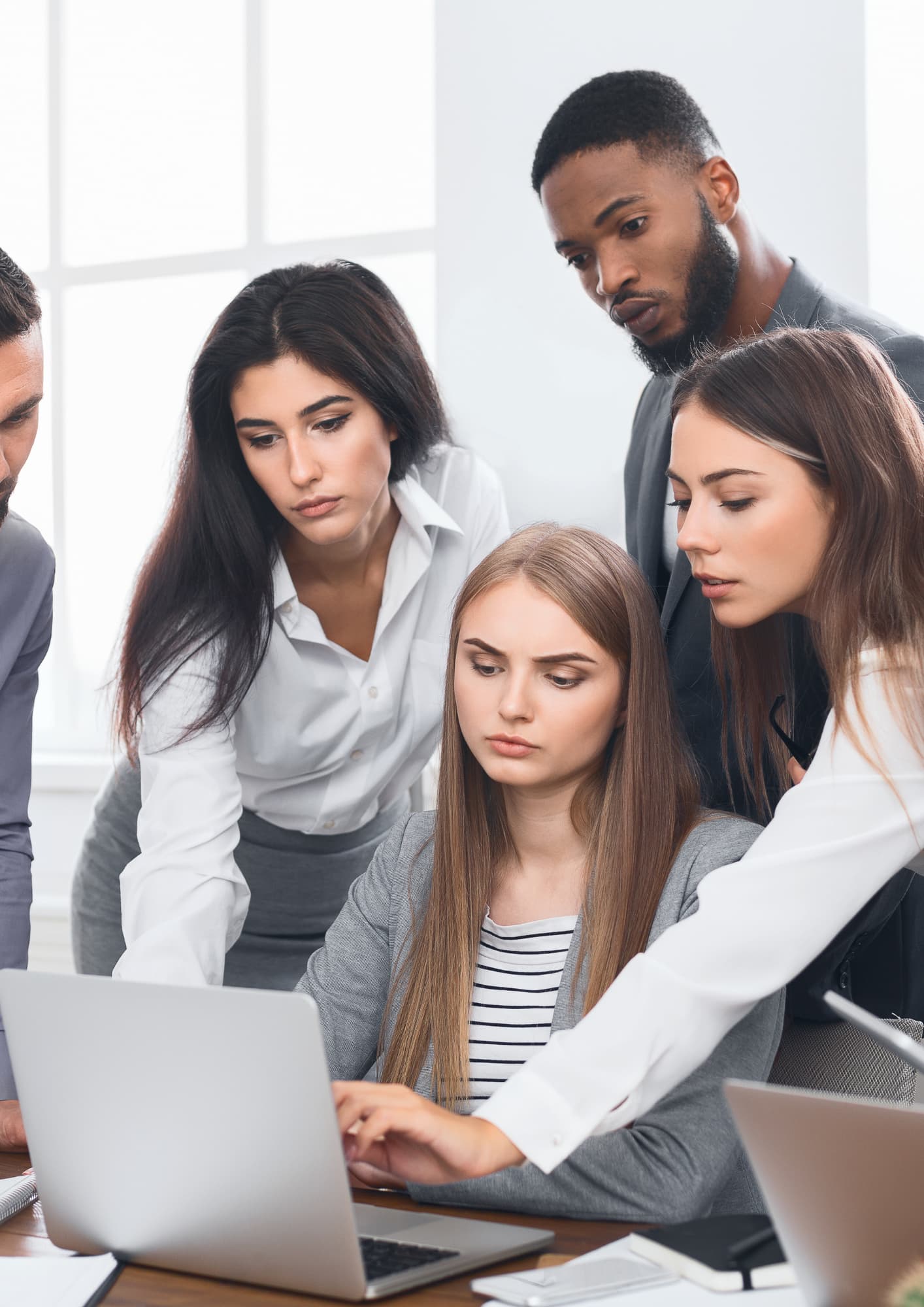 A diverse group of four professional colleagues gathering around a laptop computer in a bright office setting, looking intently at the screen during a collaborative discussion or team meeting.