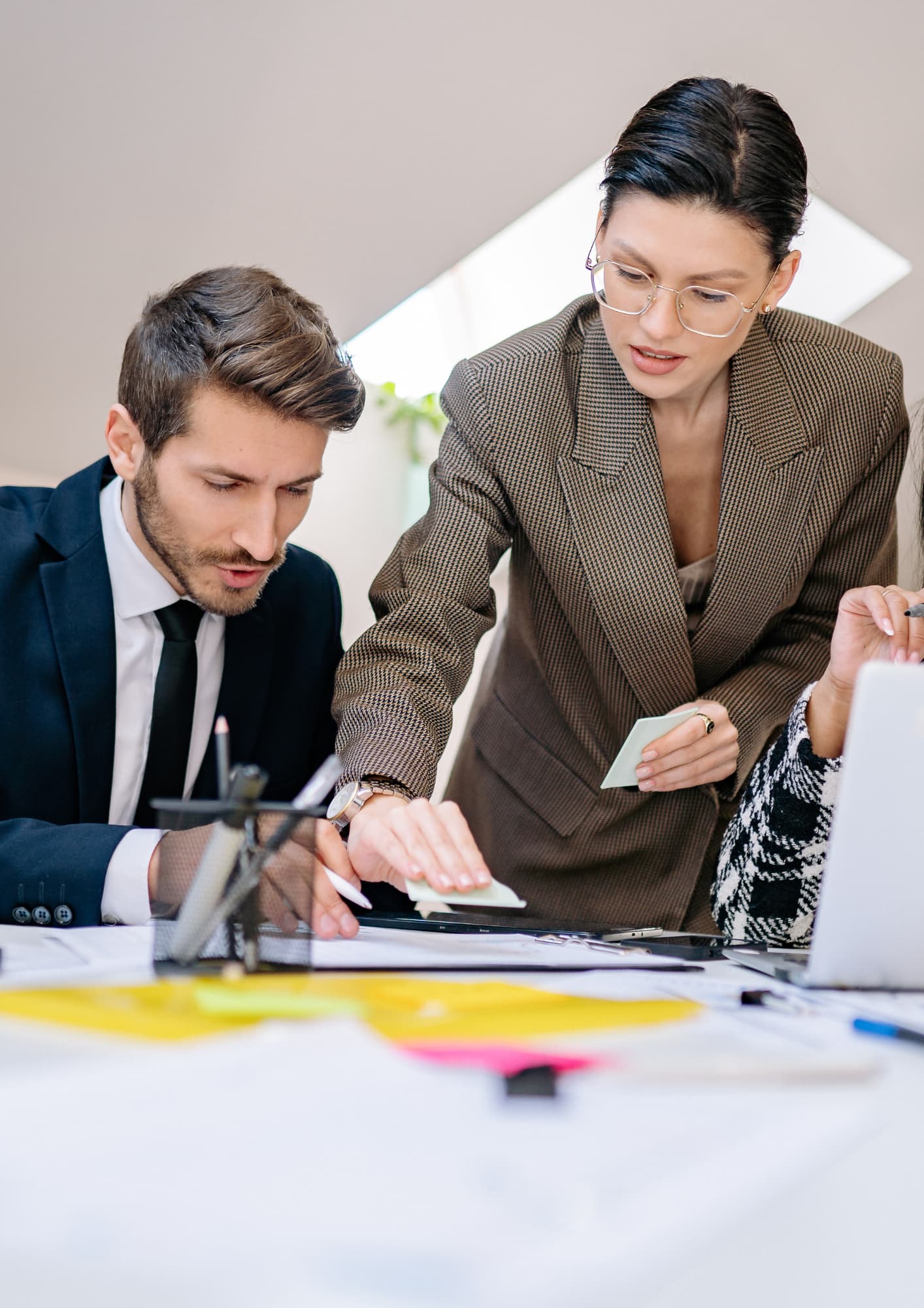 A businesswoman in a brown patterned blazer and glasses leans over a desk, holding sticky notes and pointing at a document. A businessman in a suit and tie looks down intently at the paperwork alongside her during a collaborative meeting.