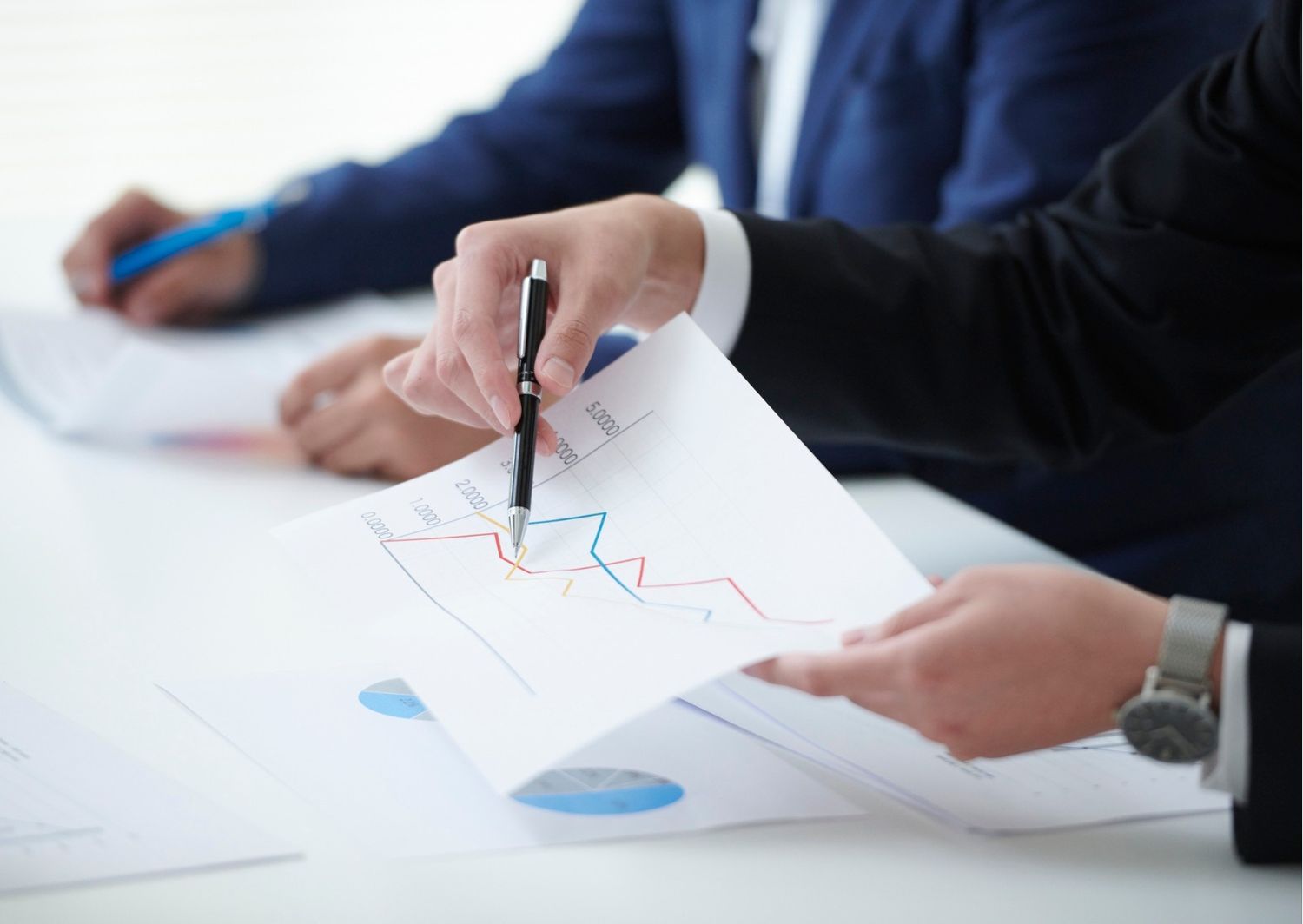 Close-up of a person's hand pointing a pen at a declining line graph on a printed document during a business meeting.  The image illustrates a downward trend in performance or sales, emphasizing the importance of identifying and addressing lost opportunities.