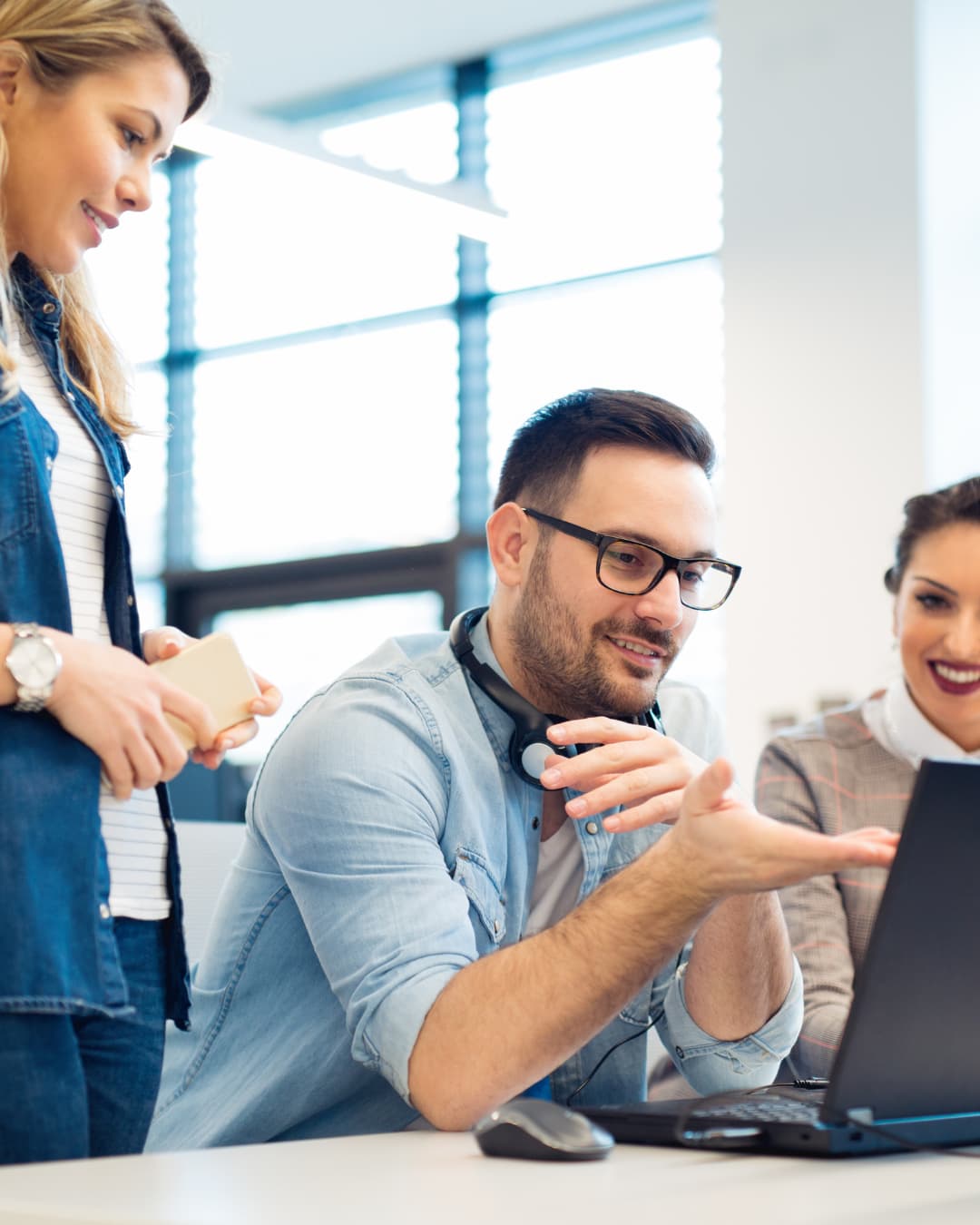 Three colleagues collaborate around a laptop in a modern office. A man wearing glasses gestures toward the
