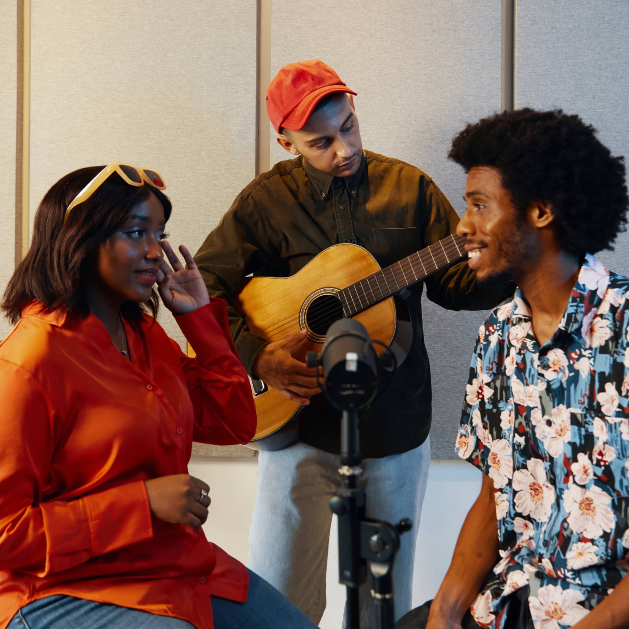 Three musicians collaborating in a studio, with one singing into a microphone while another plays acoustic guitar and a third listens and engages in discussion.