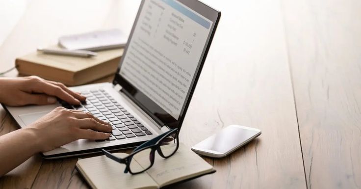 A close-up view shows a person's hands typing on a laptop keyboard, with the screen displaying a document containing text and financial data. Beside the computer on a wooden desk sit a smartphone, an open notebook holding a pair of black-rimmed glasses, and a closed book.