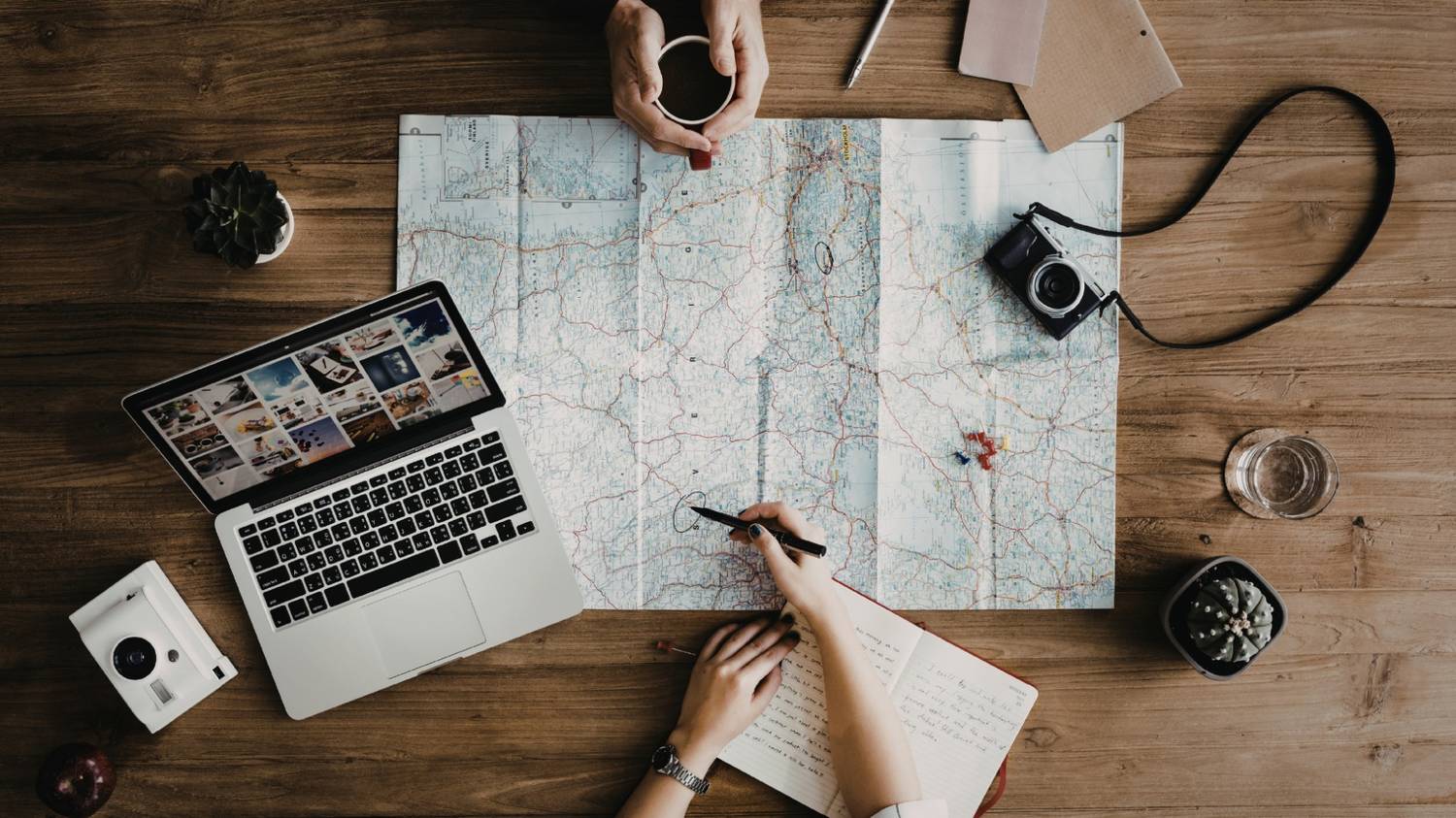 An overhead view of a wooden desk where two people are planning a route on a large paper map spread across the surface.  The scene includes a laptop, a camera, a notebook, and coffee, metaphorically representing the detailed process of mapping out a follower's journey.
