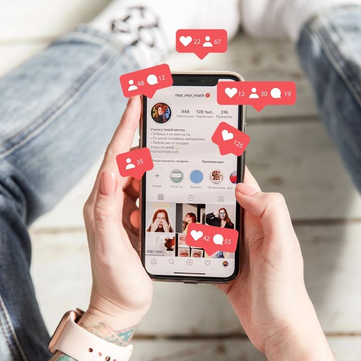 A first-person view of someone holding a smartphone that displays an Instagram profile page. Several red, 3D notification bubbles float in the air around the phone, showing white icons and numbers for likes, comments, and new followers. The person holding the phone is wearing blue jeans, patterned white socks, and a smartwatch with a light pink band.