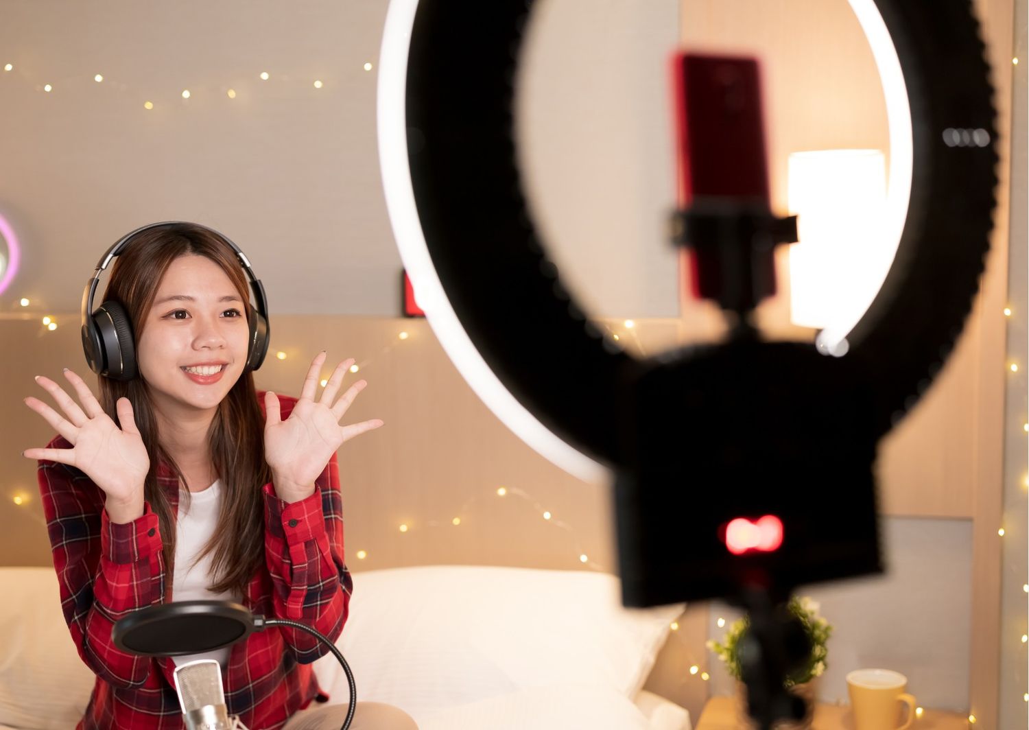 A young woman wearing headphones and a red plaid shirt smiles and waves while livestreaming into a smartphone mounted on a ring light.   The cozy indoor setting features warm fairy lights in the background, a professional microphone in the foreground, and a ring light illuminating the creator.