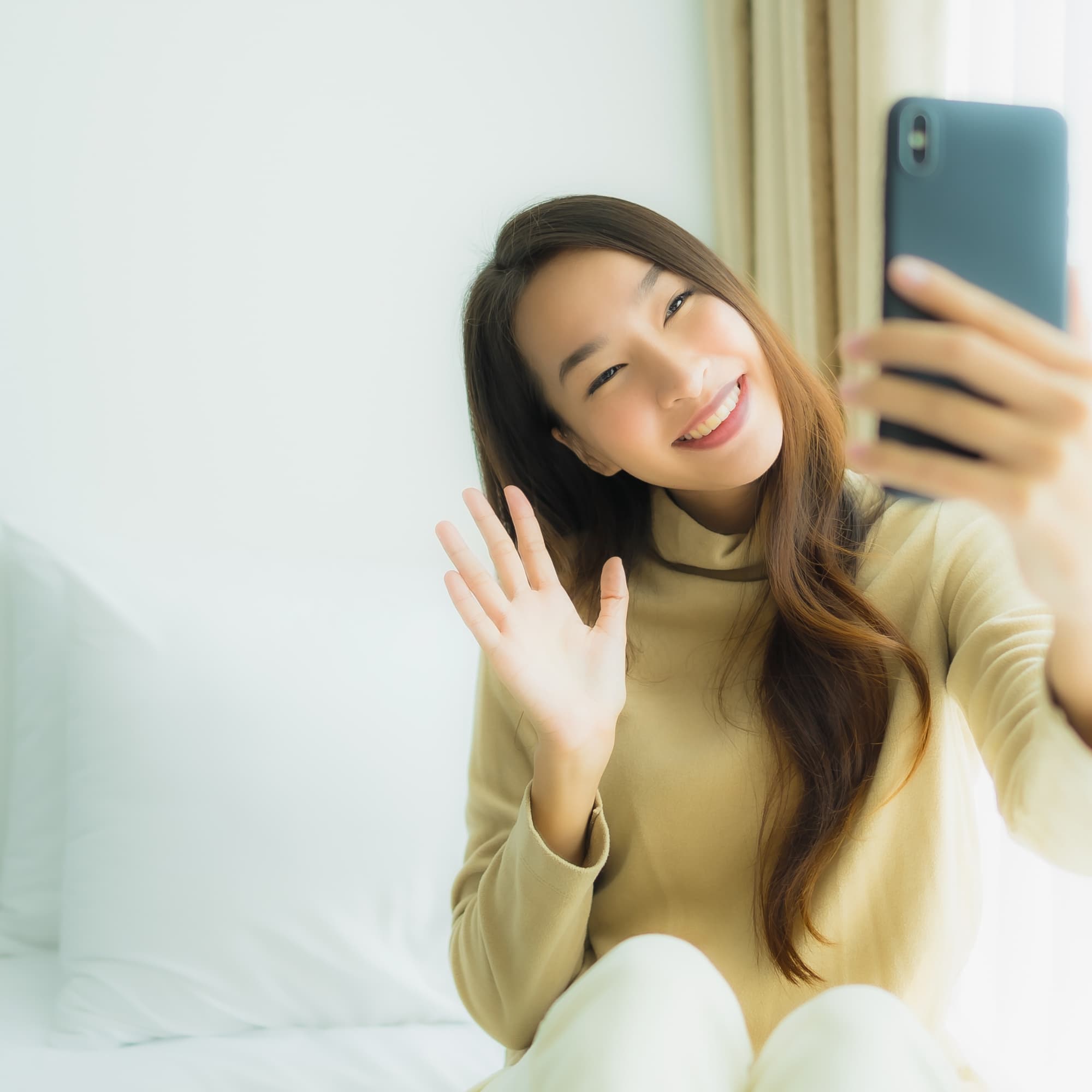 Smiling woman sitting indoors, waving while taking a selfie with her smartphone.