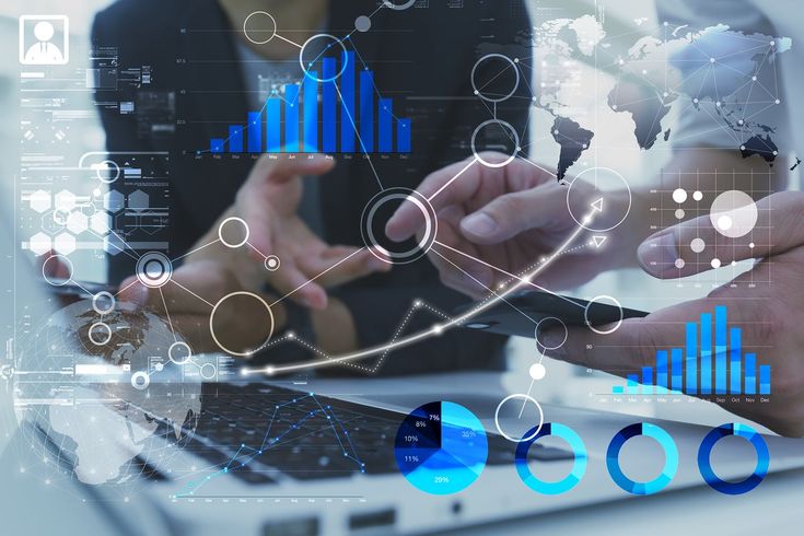 A composite digital illustration showing the hands of business professionals working with a smartphone and a laptop at a desk. The scene is heavily overlaid with complex, glowing holographic data visualizations in white and bright blue. The intricate digital graphics include rising bar charts, line graphs, interconnected network nodes, pie charts, a world map, and a glowing digital globe, conveying a strong theme of advanced data analytics, business intelligence, and global networking.