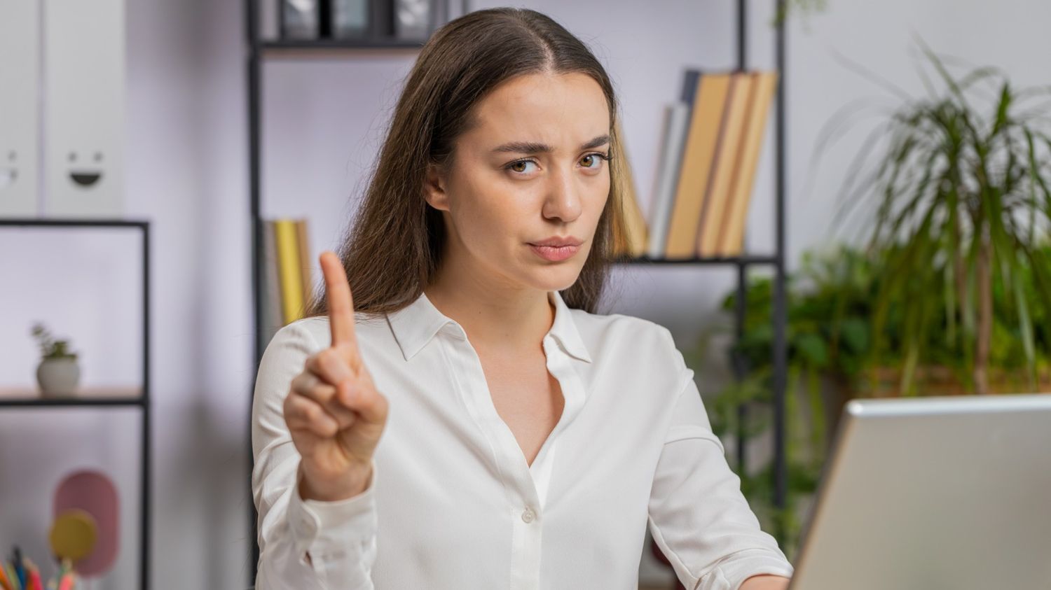 A woman sitting at a desk in an office setting, holding up one finger in a 