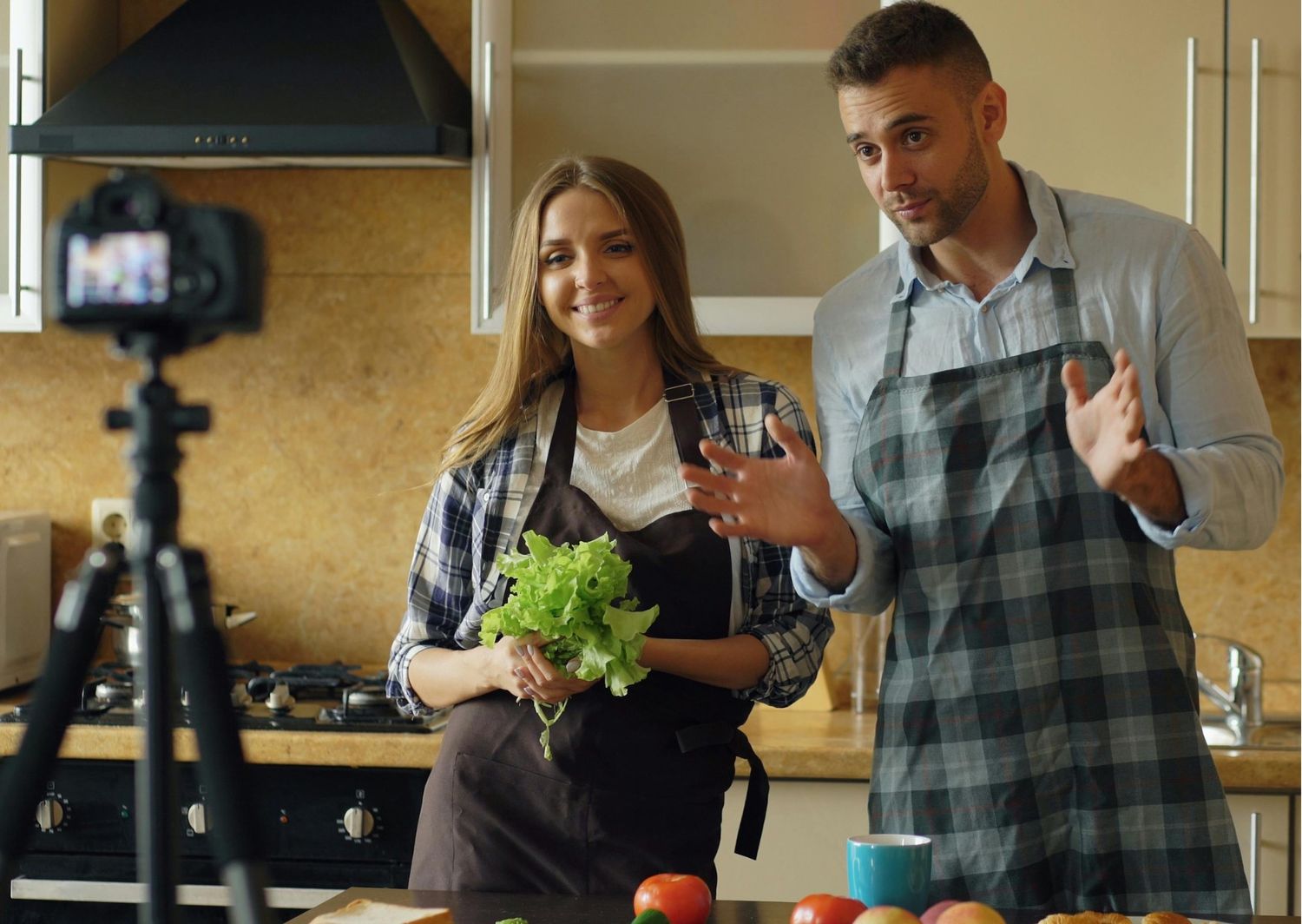 Gemini said A man and a woman, both wearing aprons, are filming a cooking or lifestyle video in a home kitchen. The camera, mounted on a tripod in the foreground, captures the duo as they present fresh ingredients like lettuce and tomatoes.