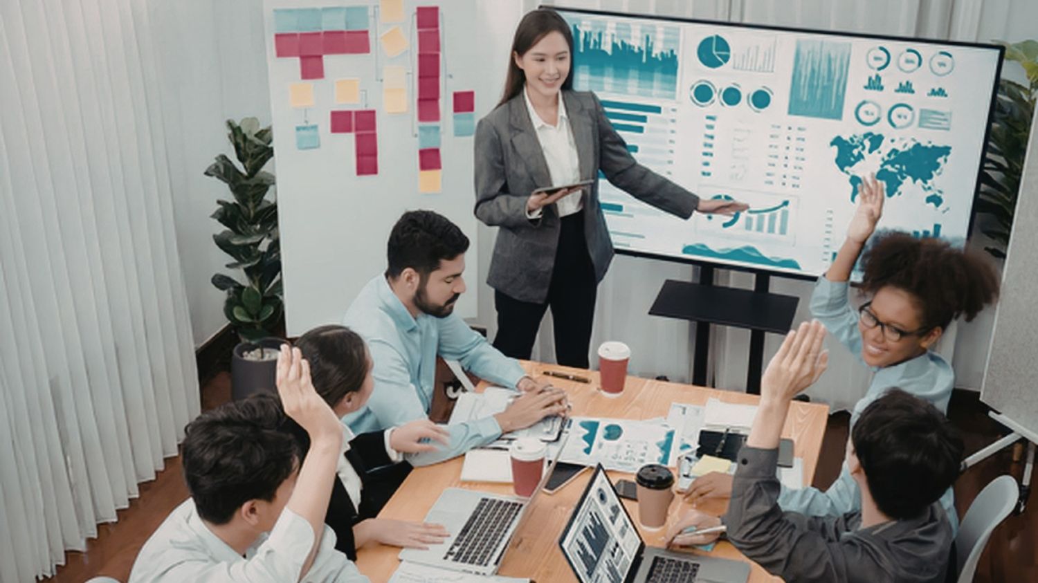 A professional woman standing by a large digital screen displaying data charts and a world map, giving a presentation to a diverse team who are raising their hands to ask questions in a modern office.