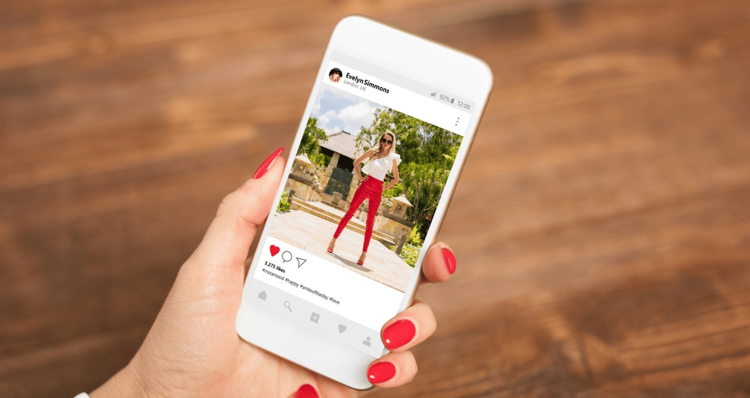 A hand with red nail polish holding a white smartphone that displays an Instagram post of a woman in a white top and red pants posing outdoors.