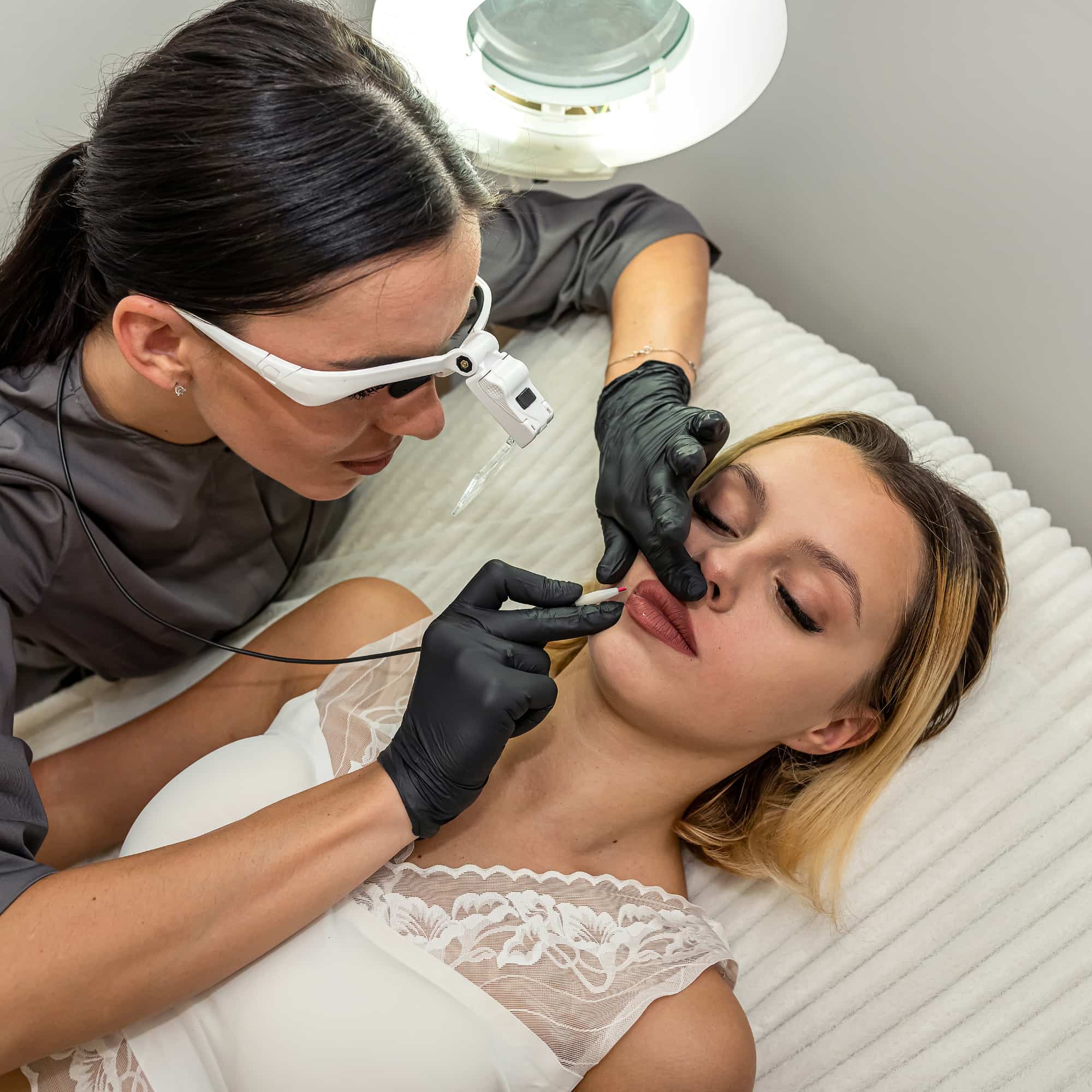 A cosmetic practitioner wearing gloves and magnifying glasses performing a lip enhancement procedure on a client in a clinical spa setting.