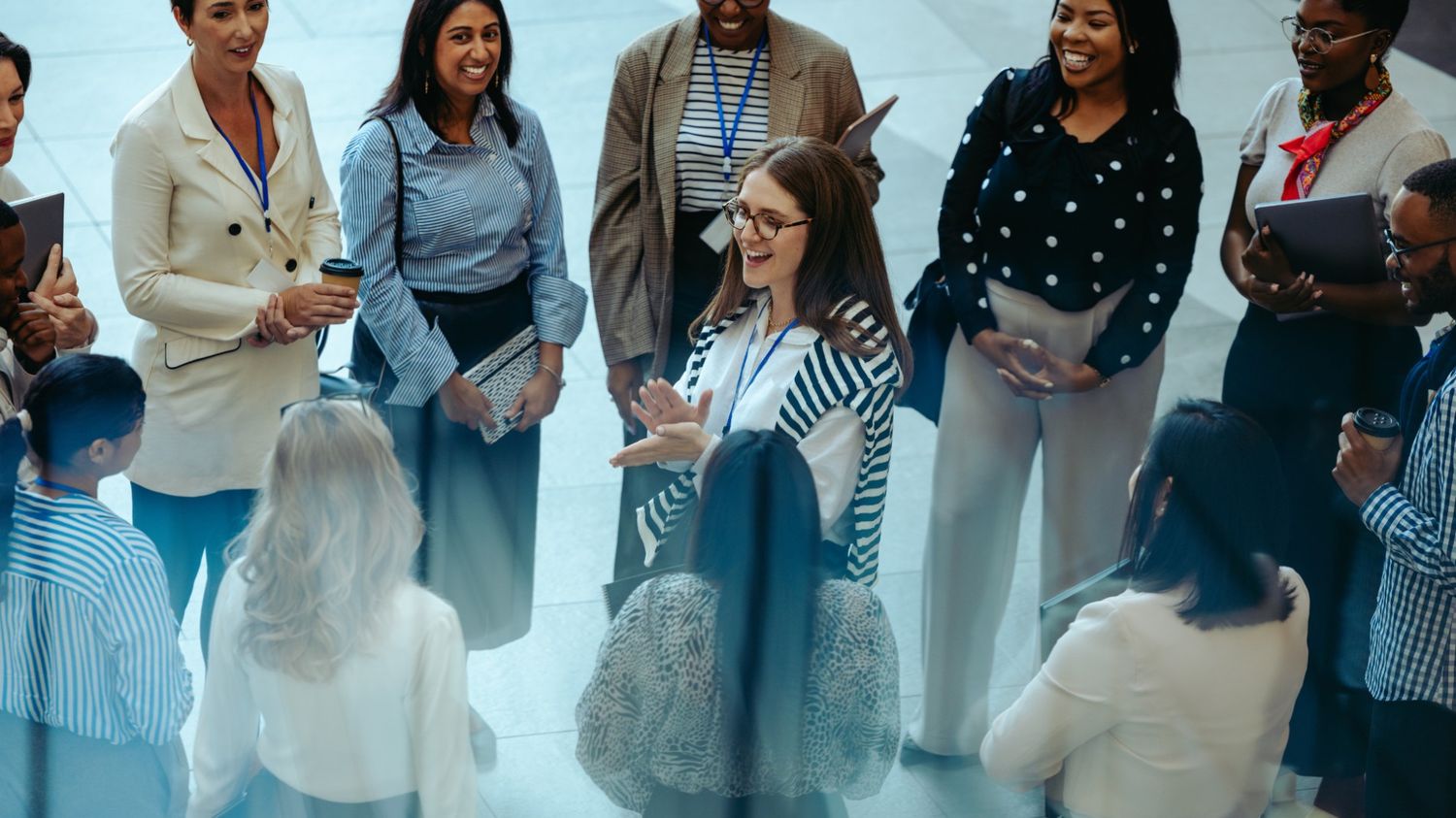 This high-angle photograph captures a diverse group of people gathered in a circle, warmly welcoming a new member. The central figure is a woman with glasses and a striped sweater draped over her shoulders, smiling and gesturing as she addresses the group.