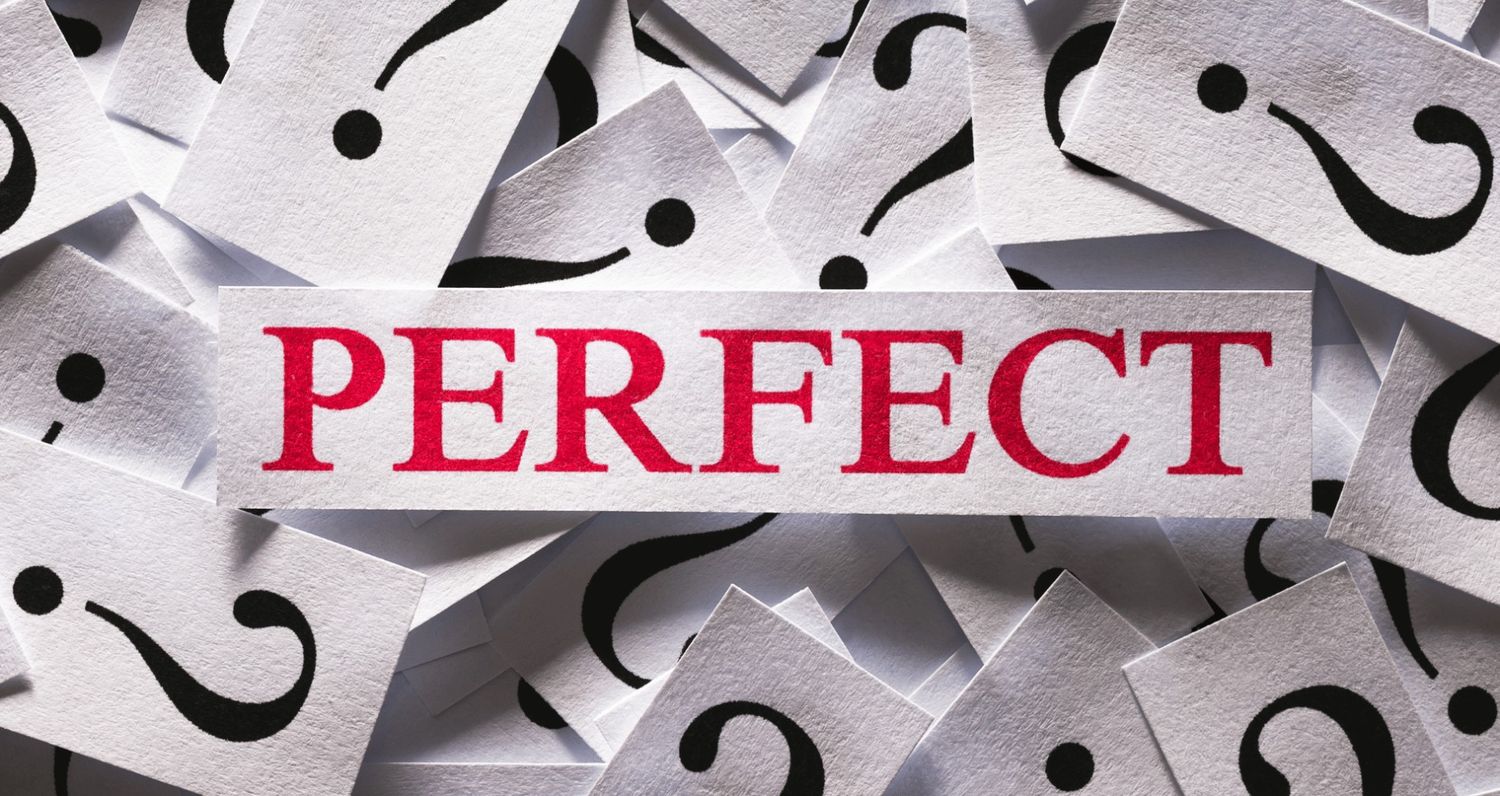 A close-up photograph of a white paper slip with the word "PERFECT" printed in bold red capital letters, placed over a scattered pile of smaller white slips featuring black question marks.