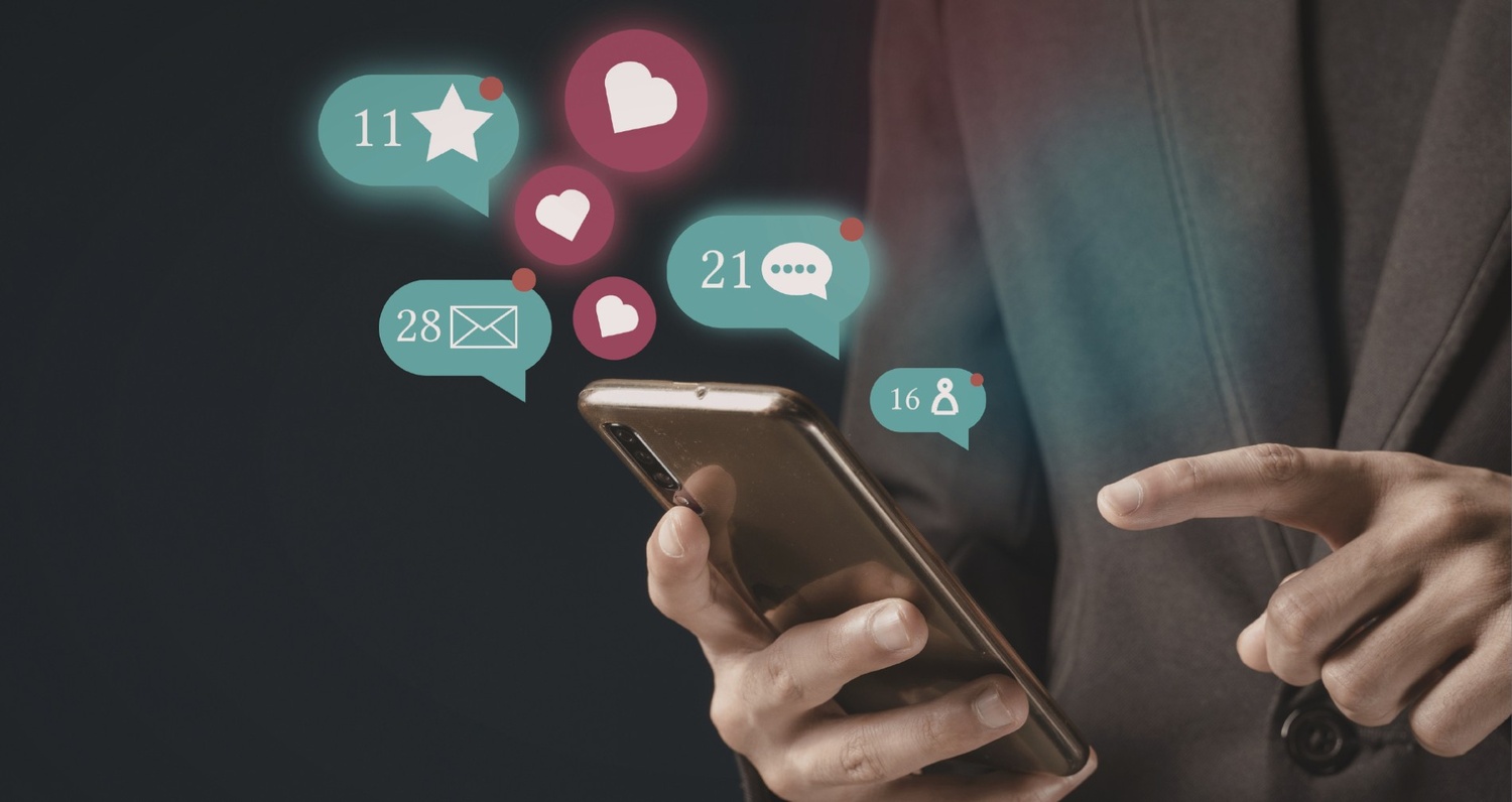 A close-up of a person in a blazer holding a smartphone with glowing social media notification icons for likes, messages, and followers floating above the screen.