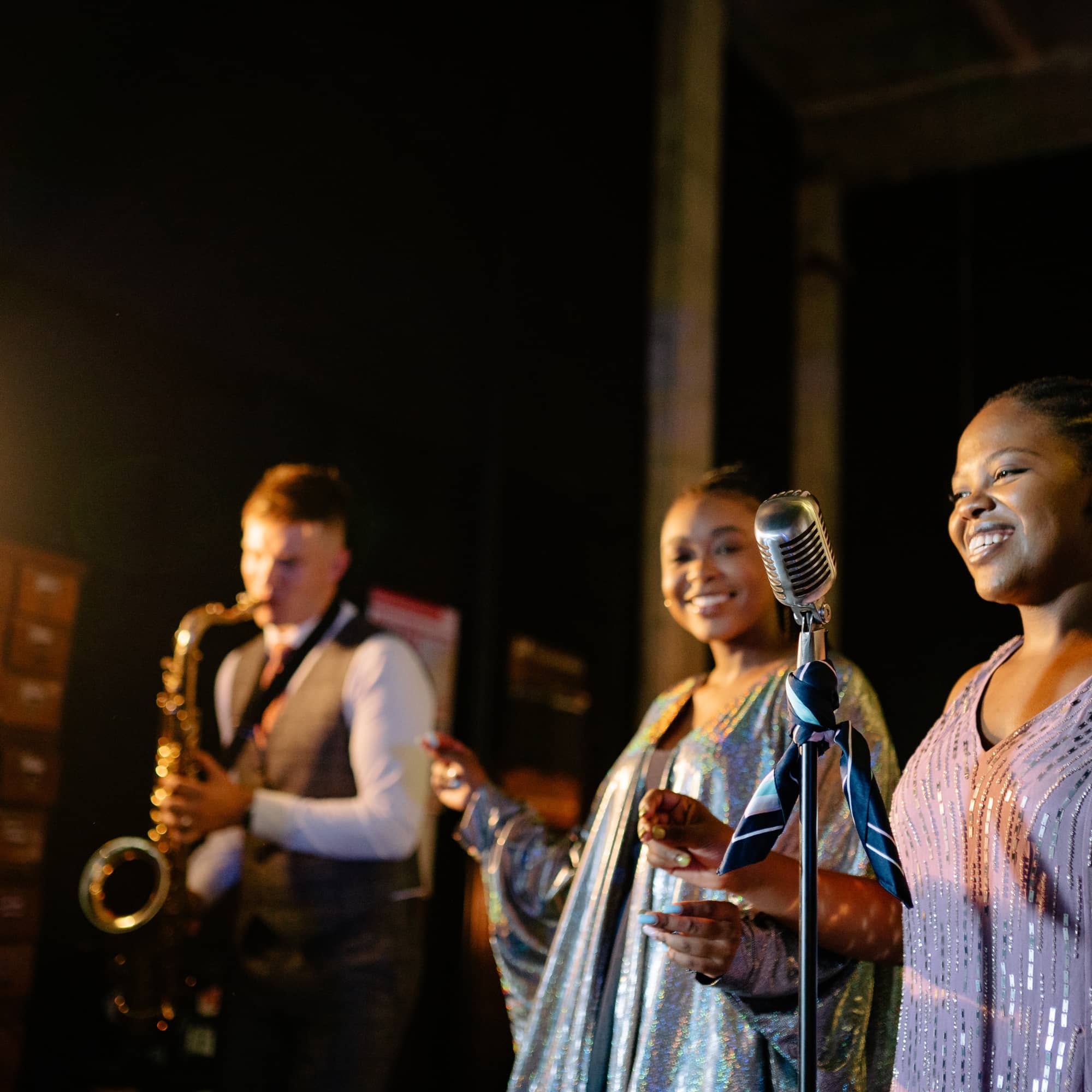 Two female singers performing on stage with a vintage microphone, smiling while a saxophonist plays in the background under warm stage lighting.