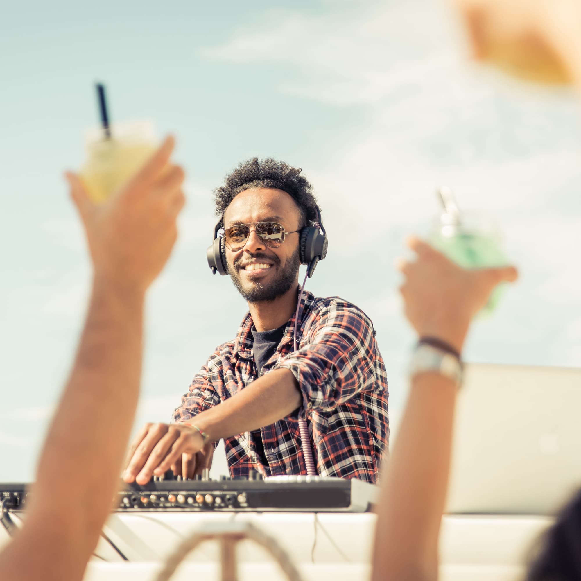 Smiling DJ wearing headphones and sunglasses performing outdoors, while people in the foreground raise drinks and enjoy the music.