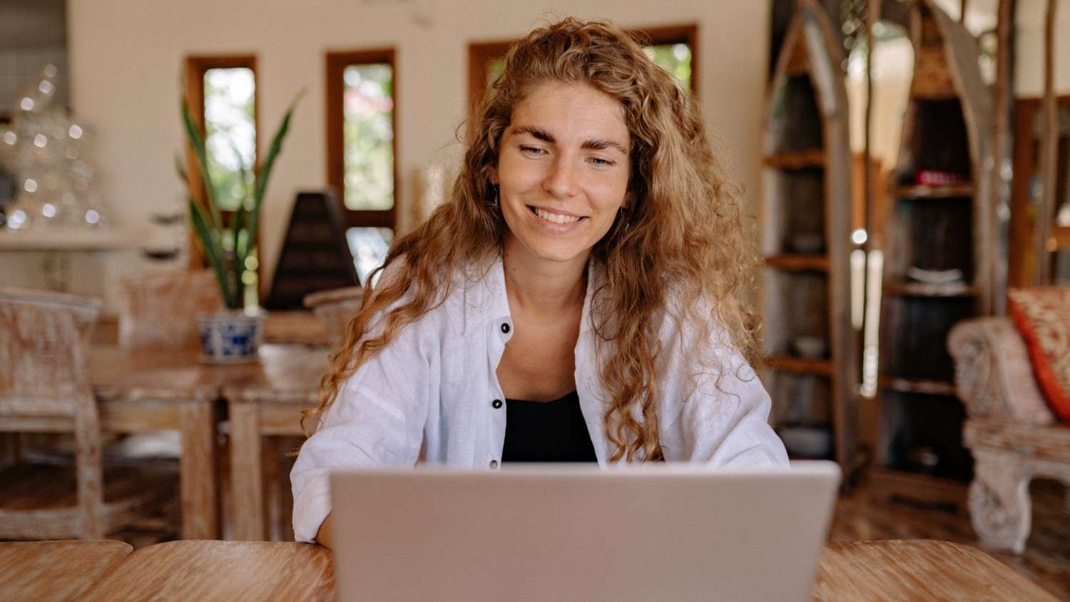 A smiling young woman with long, curly blonde hair sits at a wooden table, looking at the screen of a silver laptop. She is wearing a white button-down shirt.