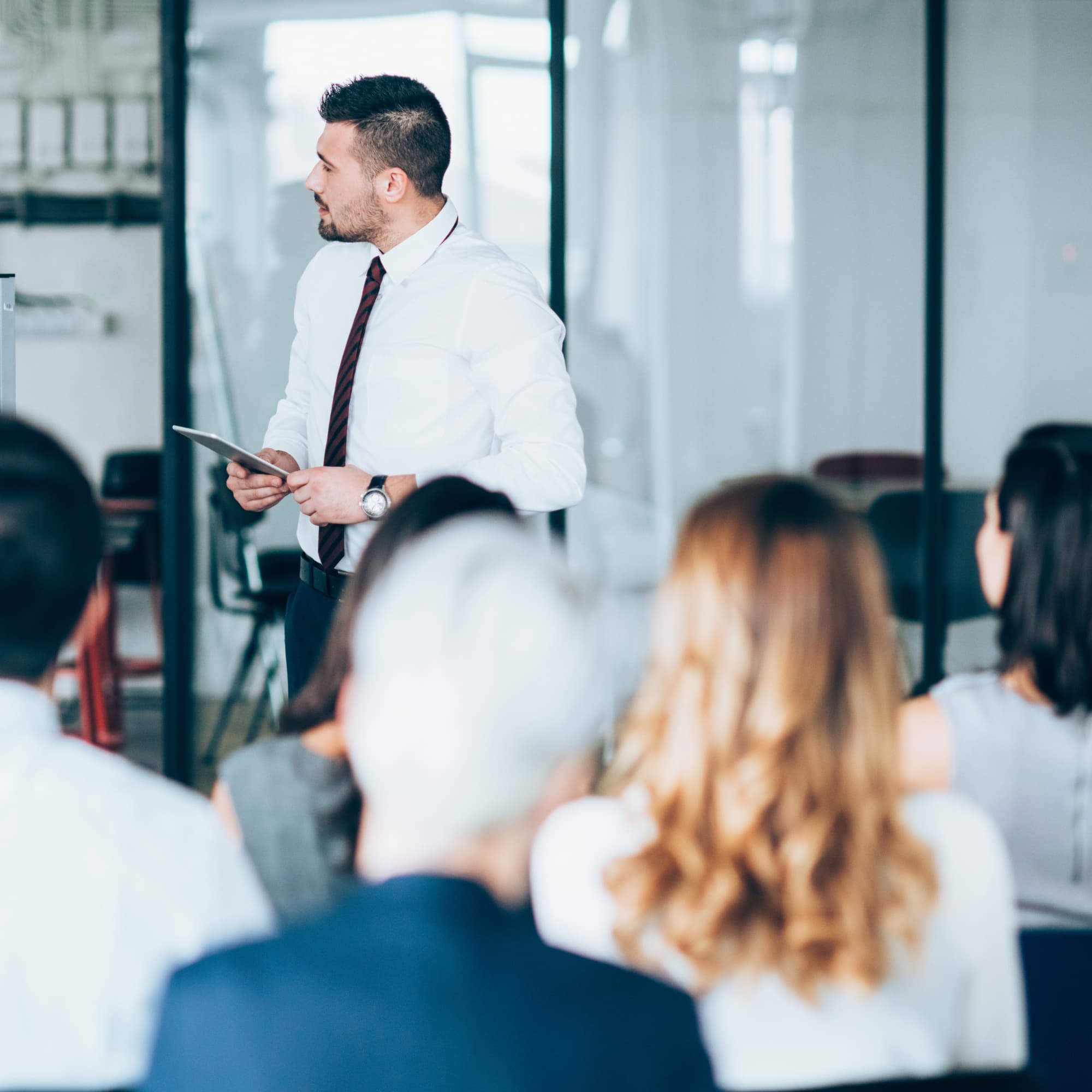 Business professional presenting to a group in a meeting room, holding a tablet while addressing an audience.