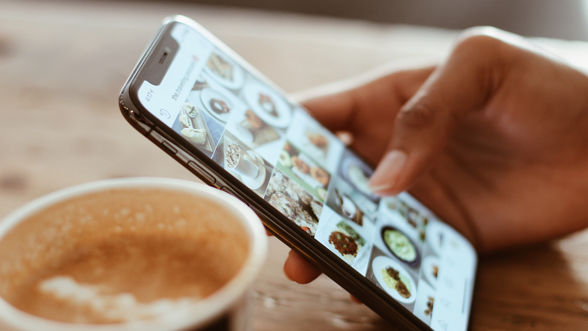 A person's hand holds a smartphone, viewing a grid of food pictures on a social media app. In the blurred foreground, a cup of frothy coffee sits on a wooden table.