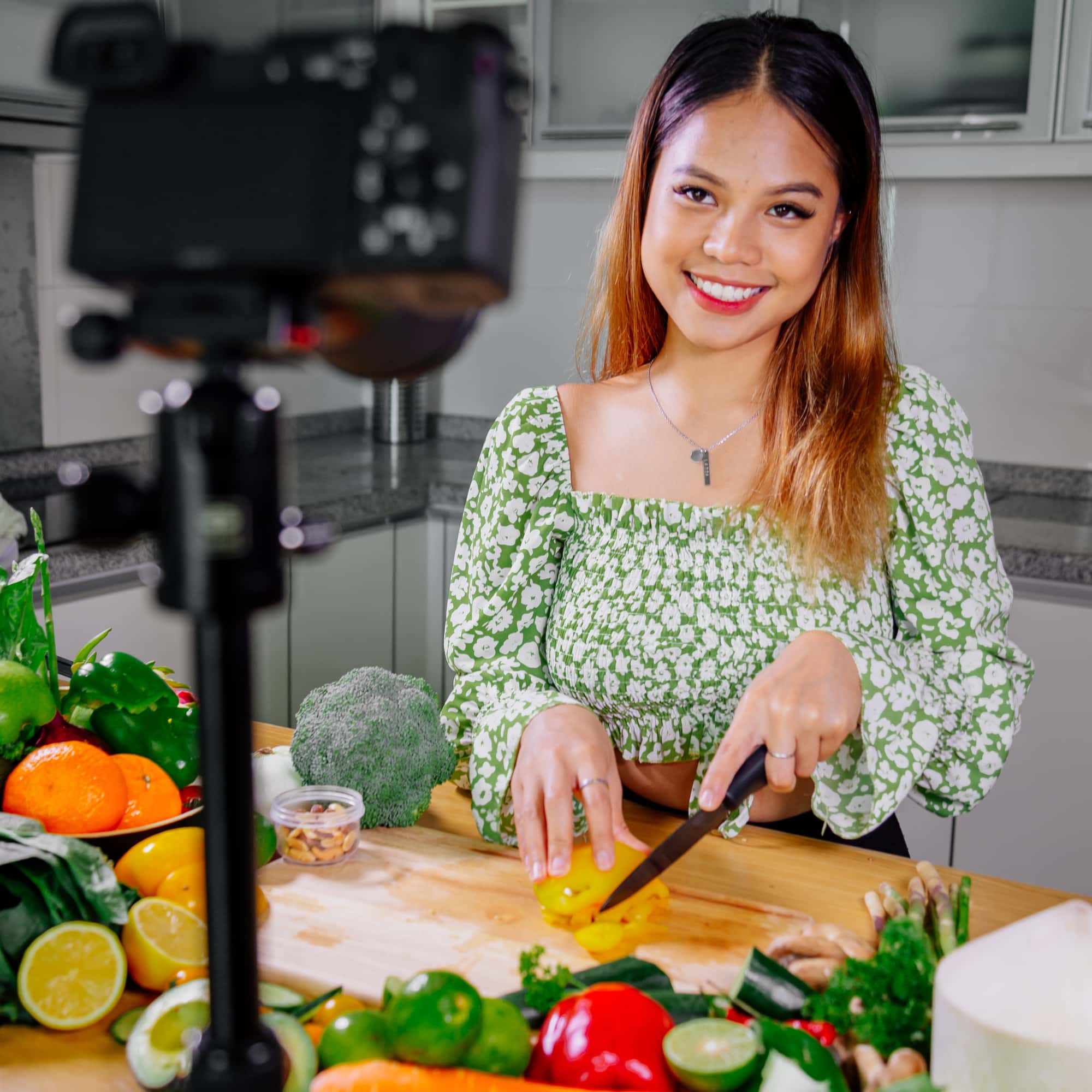 Woman smiling at a camera while chopping a yellow bell pepper on a cutting board, surrounded by fresh fruits and vegetables in a kitchen.