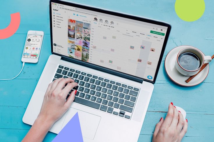 Hands typing on a laptop showing a content calendar grid, with a phone and a cup of coffee beside it on a blue desk