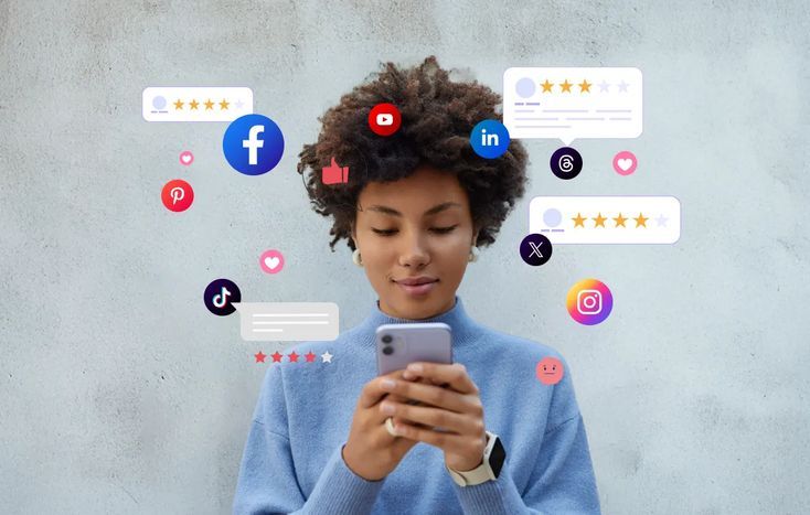 A young woman in a light blue sweater stands against a plain grey wall, looking down intently at the smartphone in her hands. Floating around her head is a digital overlay of various social media icons—including Facebook, Instagram, TikTok, and YouTube—mixed with graphic bubbles representing likes, comments, and star ratings.