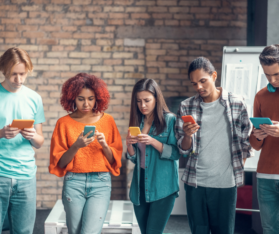 A diverse group of five young people standing in a row against a brick wall, all deeply focused on their individual smartphones and tablets.