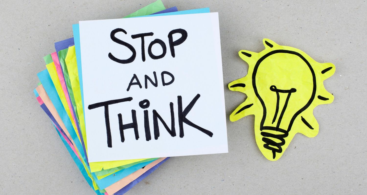 A white sticky note with the handwritten text "STOP AND THINK" in black ink, resting on a stack of colorful notes next to a yellow lightbulb cutout.