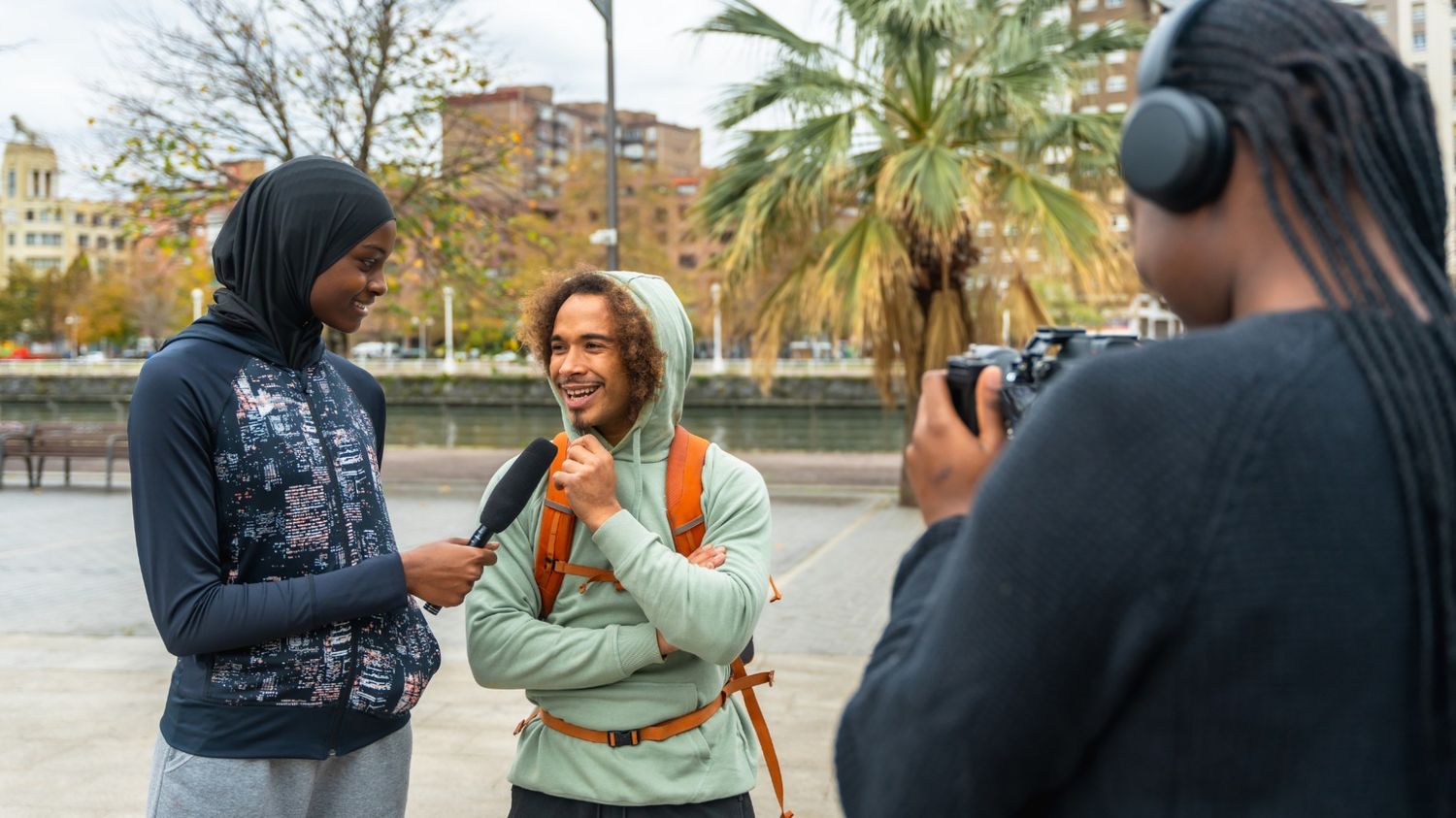 A young woman wearing a hijab holds a microphone to interview a smiling man in a hoodie outdoors, while a third person in the foreground records them with a camera and headphones.