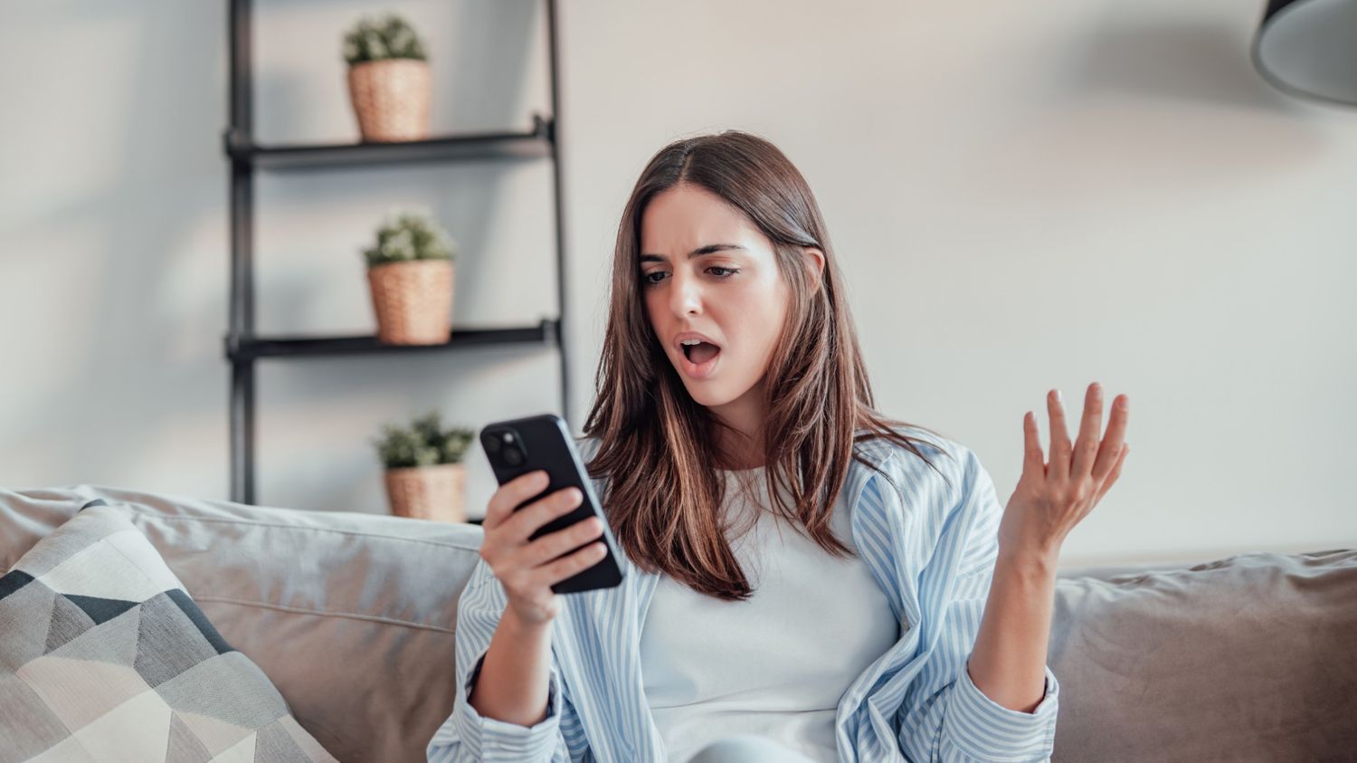 A young woman sitting on a couch, looking shocked and frustrated while staring at her smartphone.