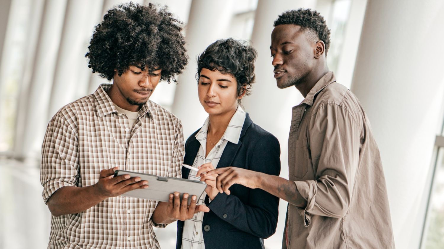 Three young professionals in a modern office hallway collaborating on a project while looking at a digital tablet held by one team member.