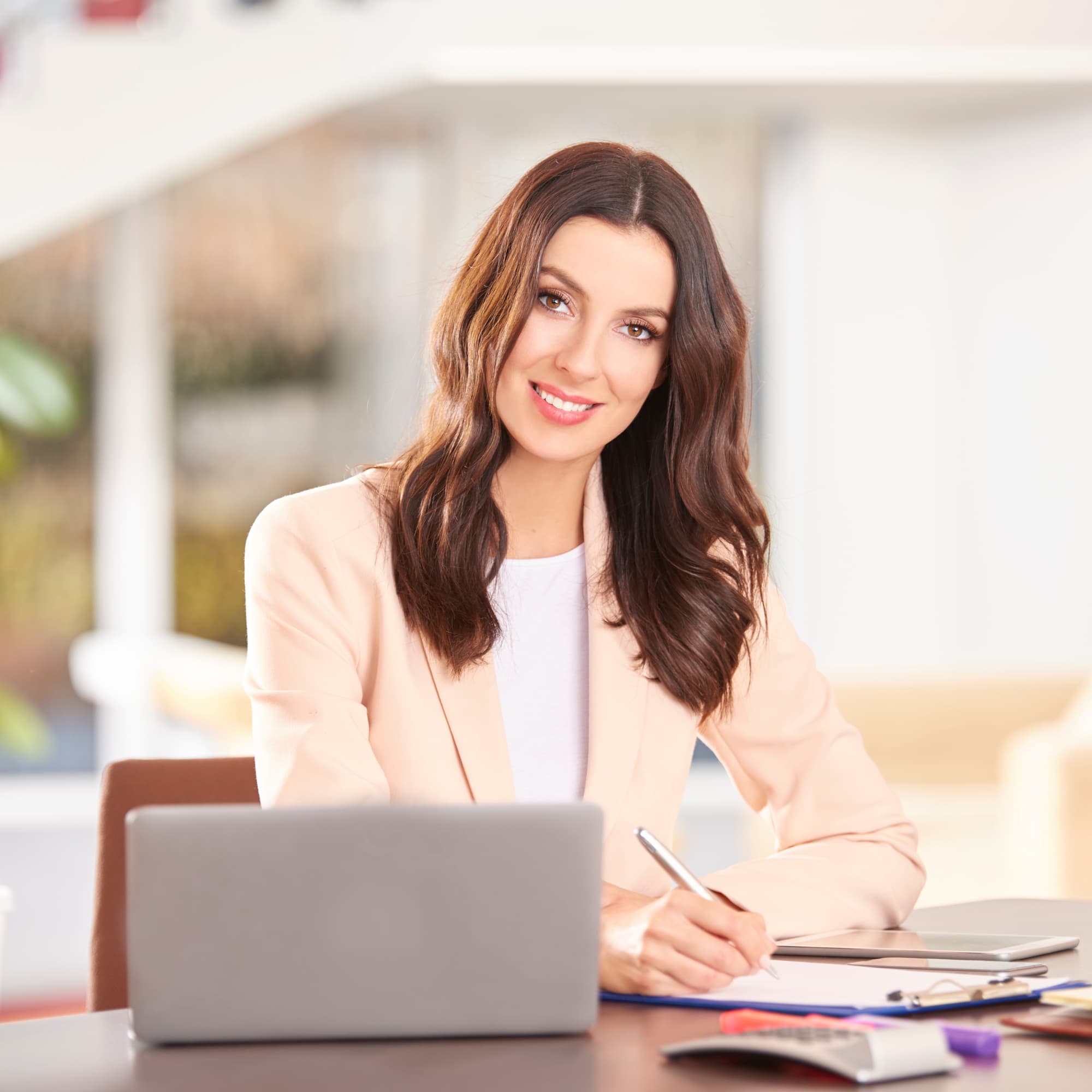 Smiling business professional sitting at a desk, writing on a clipboard while working on a laptop with a calculator nearby.