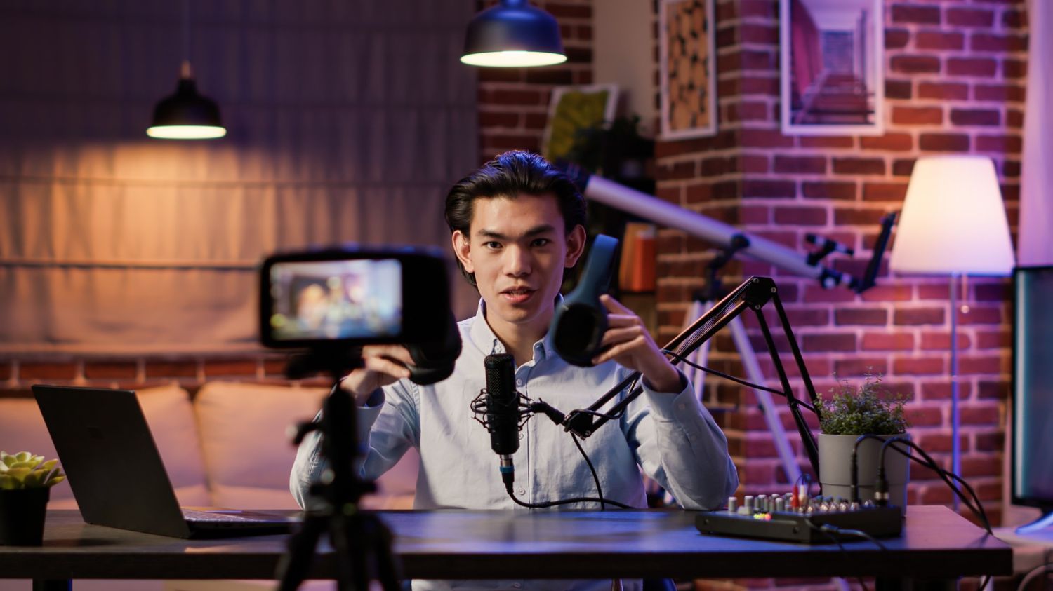 A young male creator recording a product review in a professional home studio setup. He is holding a pair of blue headphones toward a camera on a tripod, with a dedicated microphone, audio mixer, and warm ambient lighting in the background, representing high-quality broadcast and community engagement.