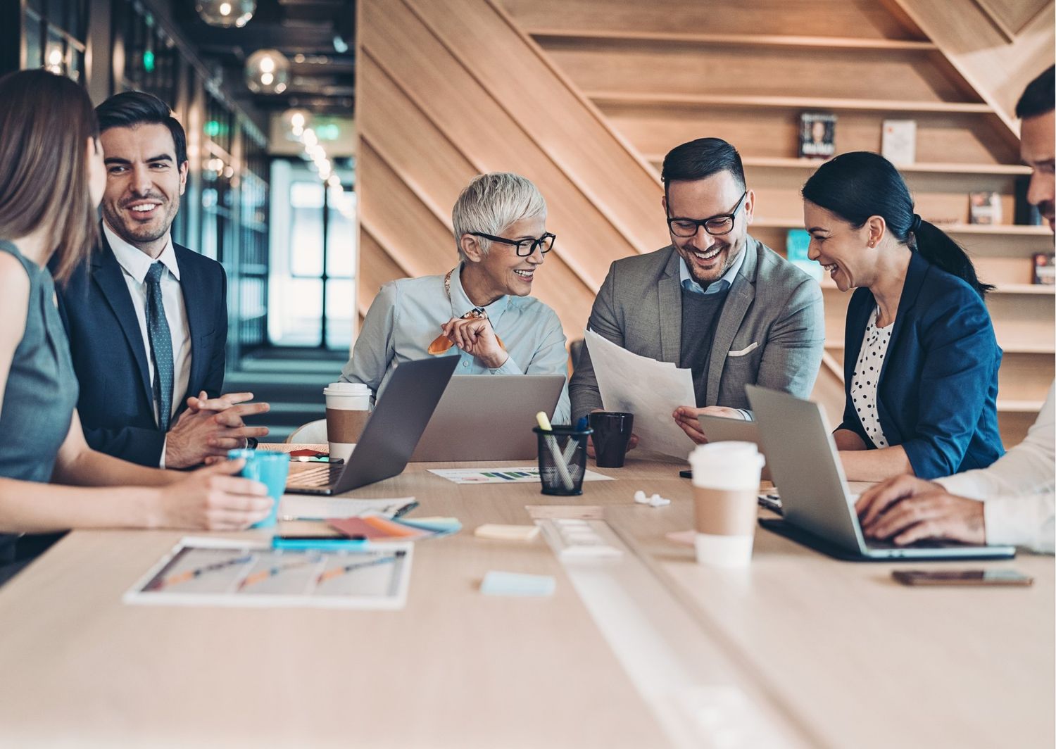 A professional, candid photograph of a diverse group of five colleagues sitting around a conference table in a modern office, laughing and engaged in a collaborative discussion over documents and laptops.