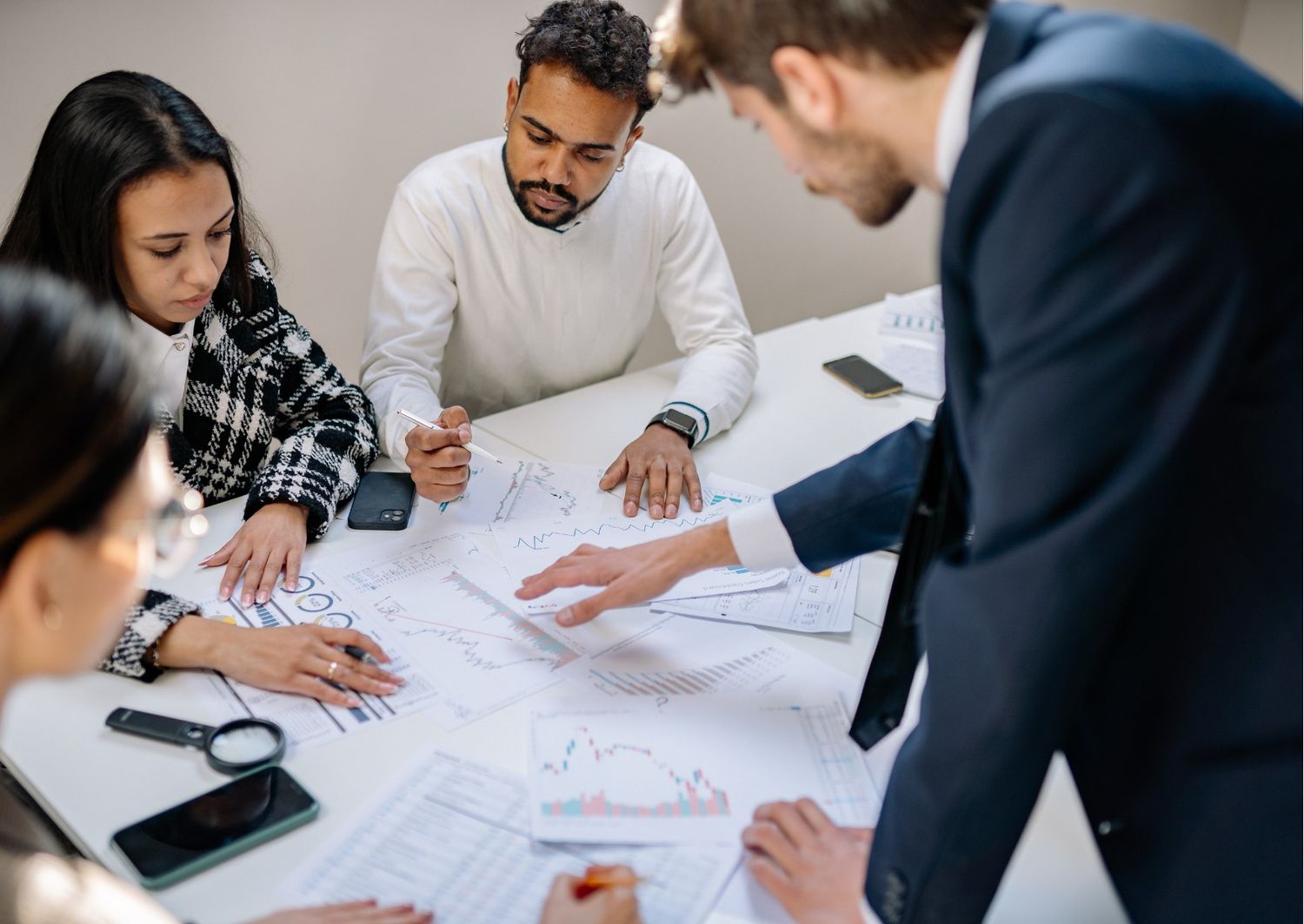 A group of professionals is gathered around a white desk, deeply engaged in analyzing a variety of printed data charts and financial graphs. One person points toward a specific trend on a document, leading a collaborative discussion with colleagues who are focused and taking notes.  This scene illustrates the **analytical and psychological depth** behind marketing strategies. It emphasizes that successful "comment-to-DM" funnels are built on data-driven insights, careful planning, and a thorough understanding of consumer behavior.