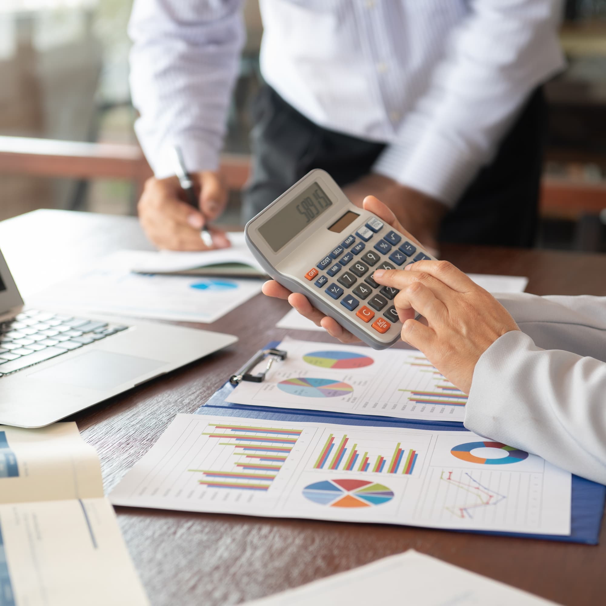 Close-up of hands using a calculator over financial reports with charts and graphs, while another person writes in the background near a laptop.