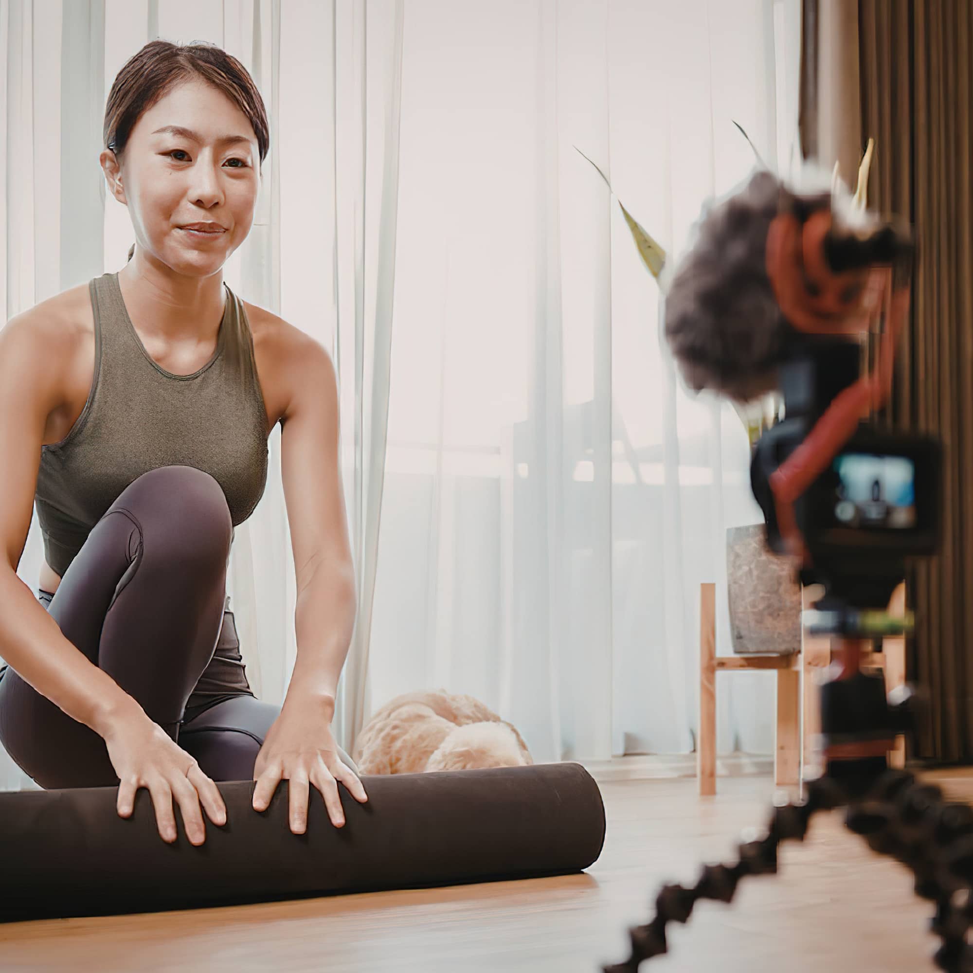 Woman in activewear sitting on a yoga mat, stretching and recording a fitness video with a camera on a tripod at home.