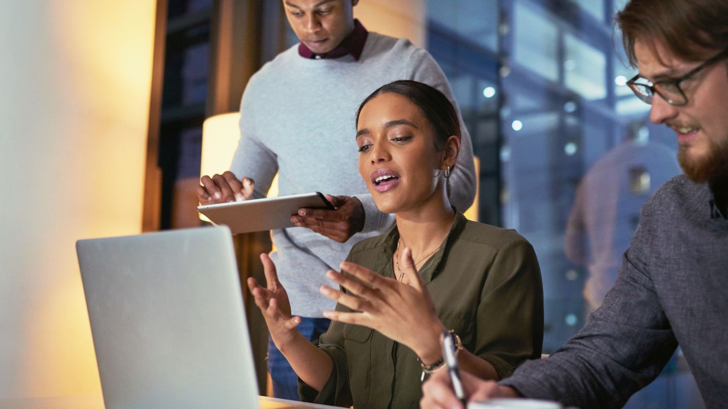 This image captures a collaborative moment in a modern office, featuring a woman mid-explanation while gesturing toward a laptop. A colleague stands behind her with a tablet, and another sits beside her taking notes, all deeply focused on the project at hand.  Continuing the strategic journey of your digital marketing series, this visual represents the "Innovation & Adaptation" phase—the internal shift required to keep up with an platform like Instagram.