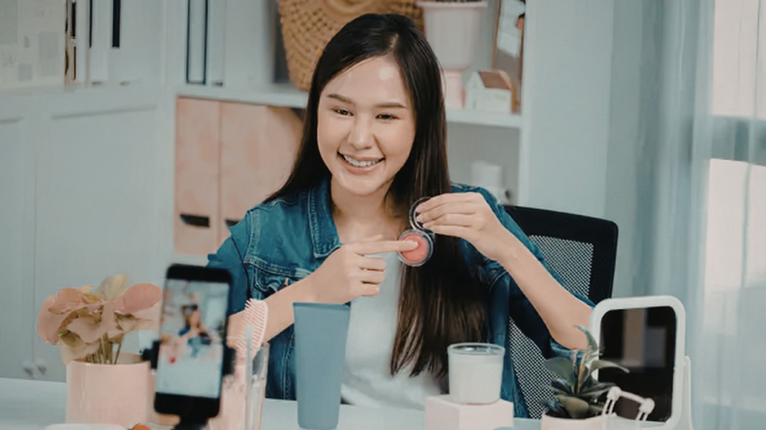 A smiling female creator in a denim jacket sits at a desk, demonstrating a makeup product to a smartphone camera mounted on a tripod for a social media video.