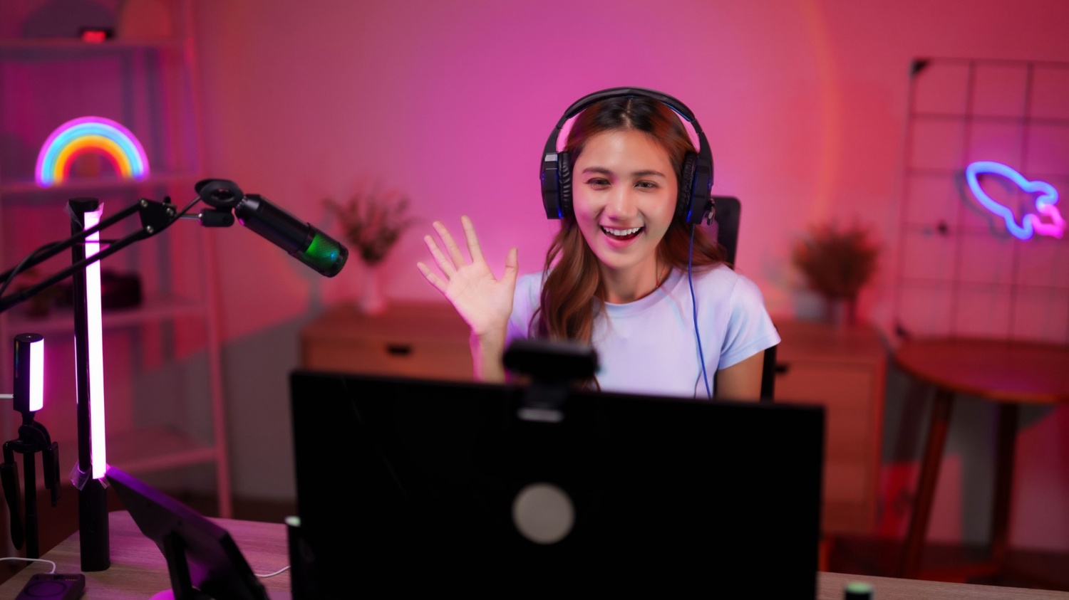 A creator wearing headphones waves at a webcam in a neon-lit streaming room, with a microphone and ring lights around the desk