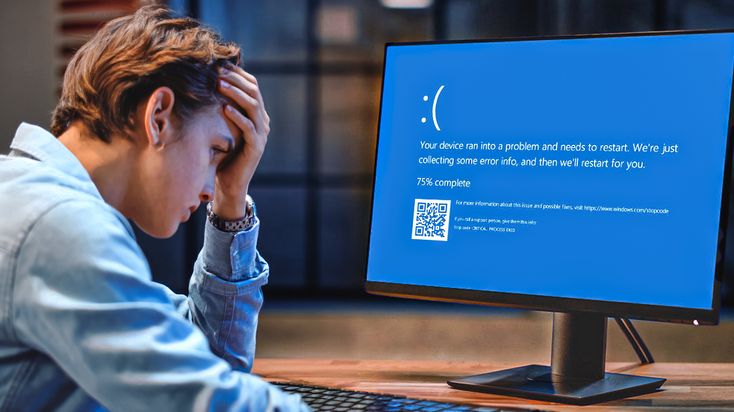 A frustrated young man in a blue denim shirt sits at a desk, clutching his head while staring at a computer monitor. The screen displays a bright blue 