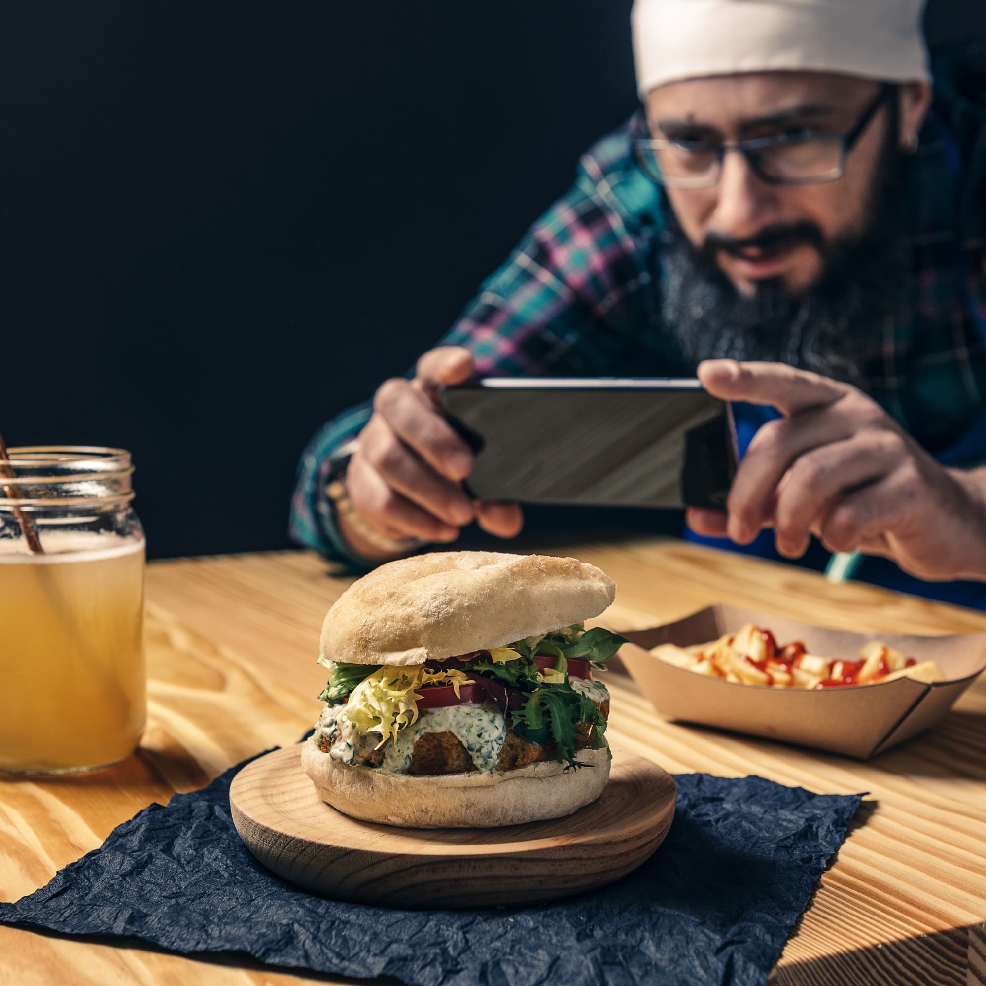 Man wearing glasses and a headband photographing a burger with his smartphone, with fries and a drink on the table.