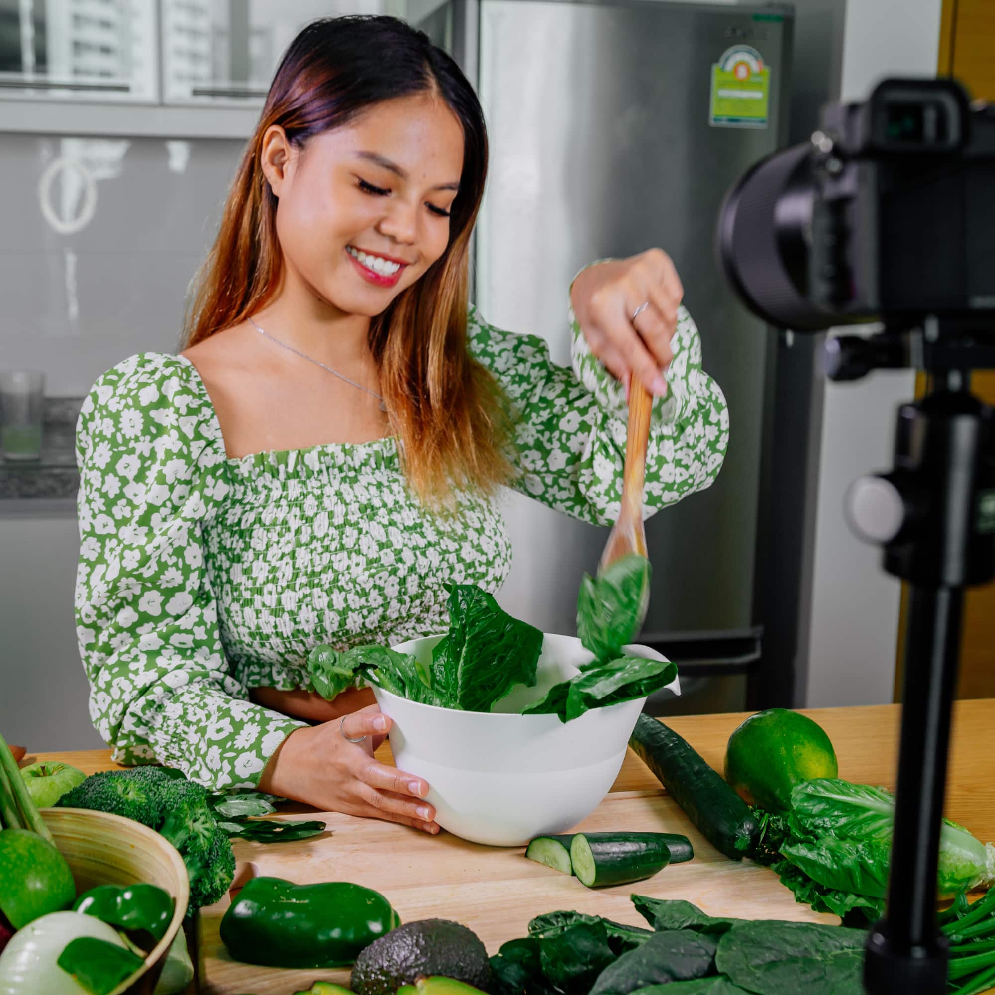 Woman mixing a green salad in a bowl while recording with a camera in a kitchen, surrounded by fresh vegetables.