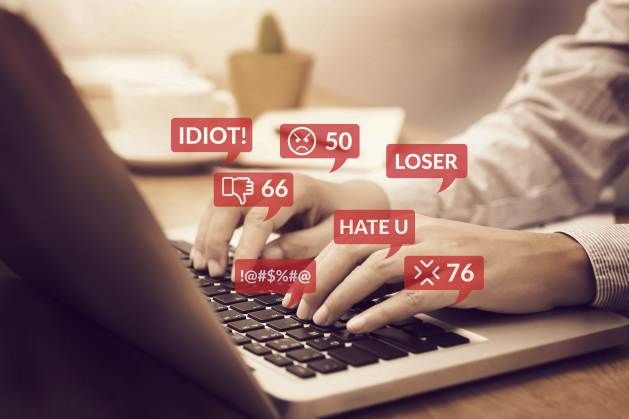 A close-up of hands typing on a laptop keyboard, surrounded by floating red speech bubbles containing negative comments and angry emojis.