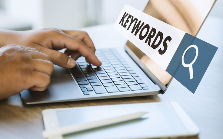 A close-up of a person's hands typing on a silver laptop keyboard on a wooden desk. A graphic overlay of a search bar containing the word