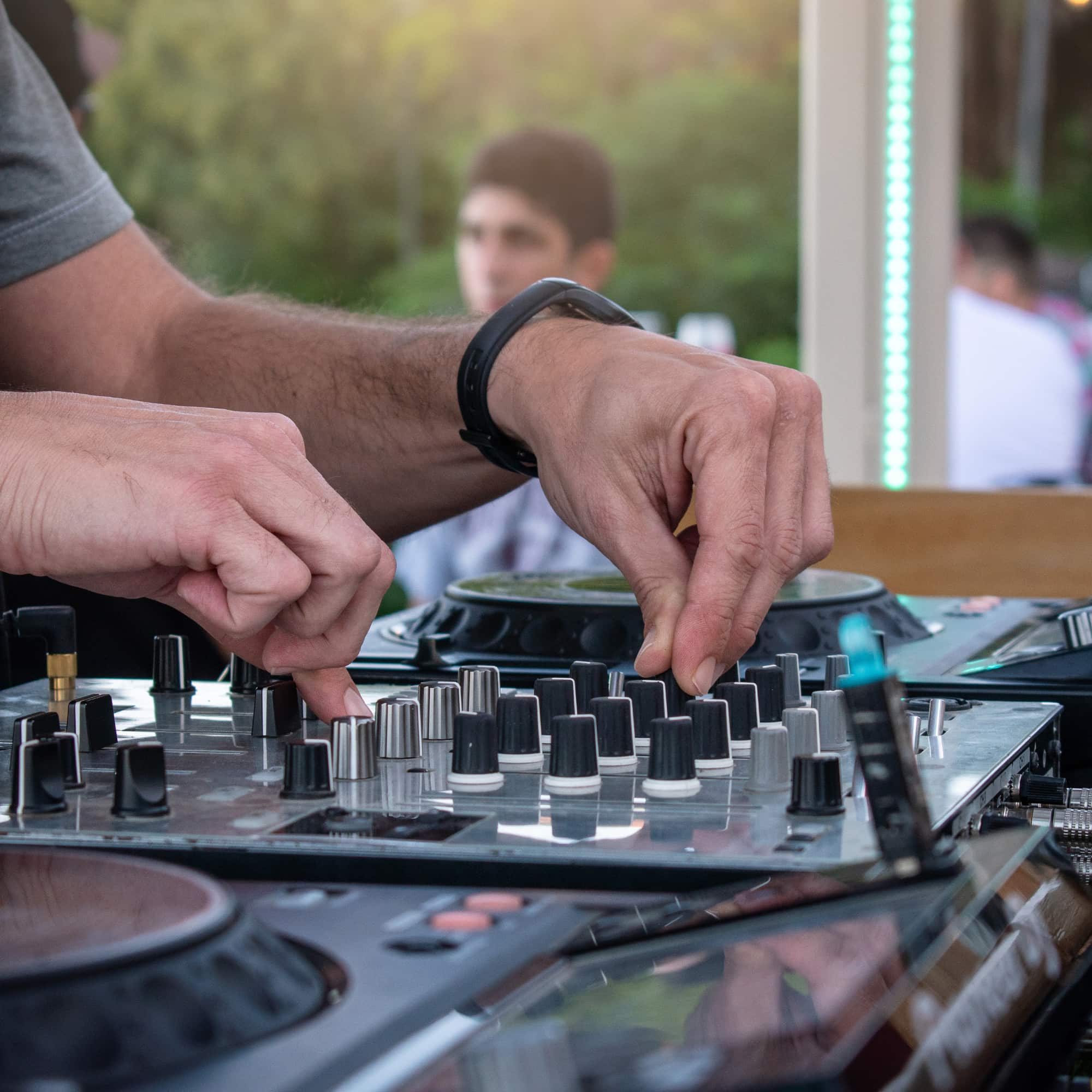 Close-up of a DJ’s hands adjusting knobs and controls on a mixing console at an outdoor event, with blurred people and greenery in the background.
